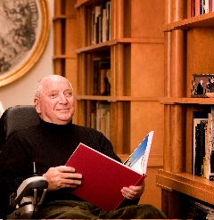 A smiling person in a black turtleneck sits in a wheelchair, holding an open red book in a library with wooden shelves.