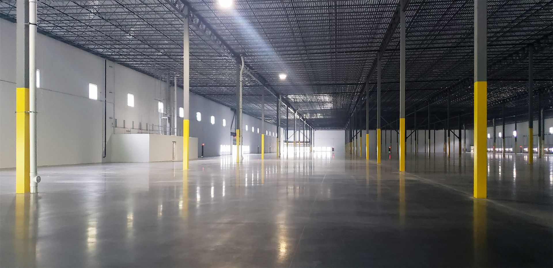 A spacious, empty industrial warehouse with polished concrete floors, tall support pillars with yellow bases, and a high ceiling.