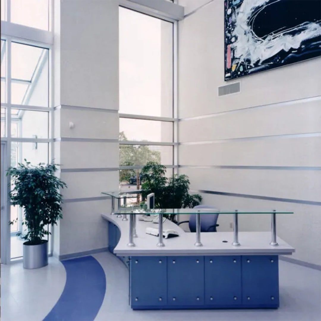 A bright, modern lobby with a blue curved reception desk, tall windows, and potted plants against white walls.