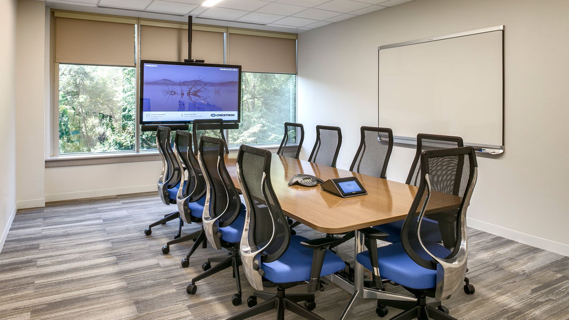 A modern conference room with a wooden table, blue office chairs, a flat-screen TV, and a blank whiteboard on the wall.
