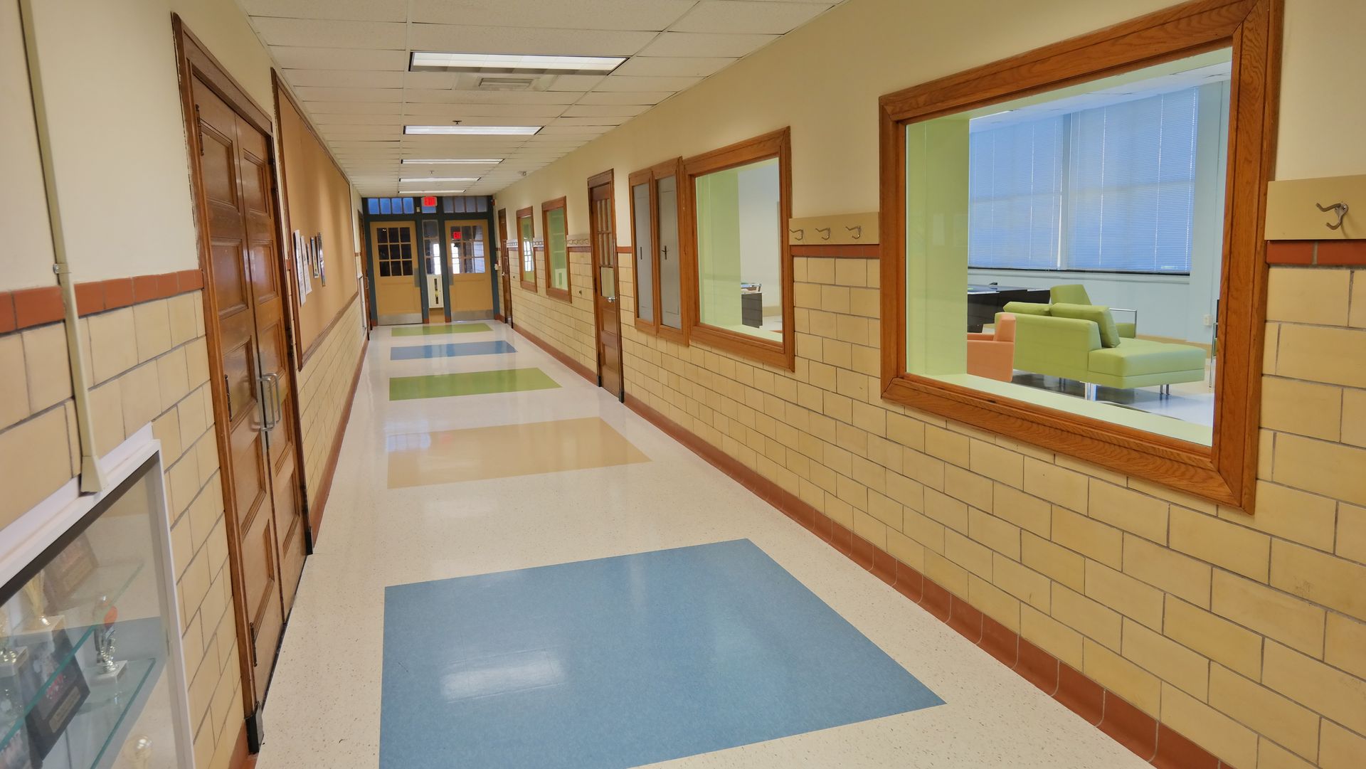 A long, empty school hallway with tan brick walls, wood-framed windows, and colorful rectangular floor tiles.