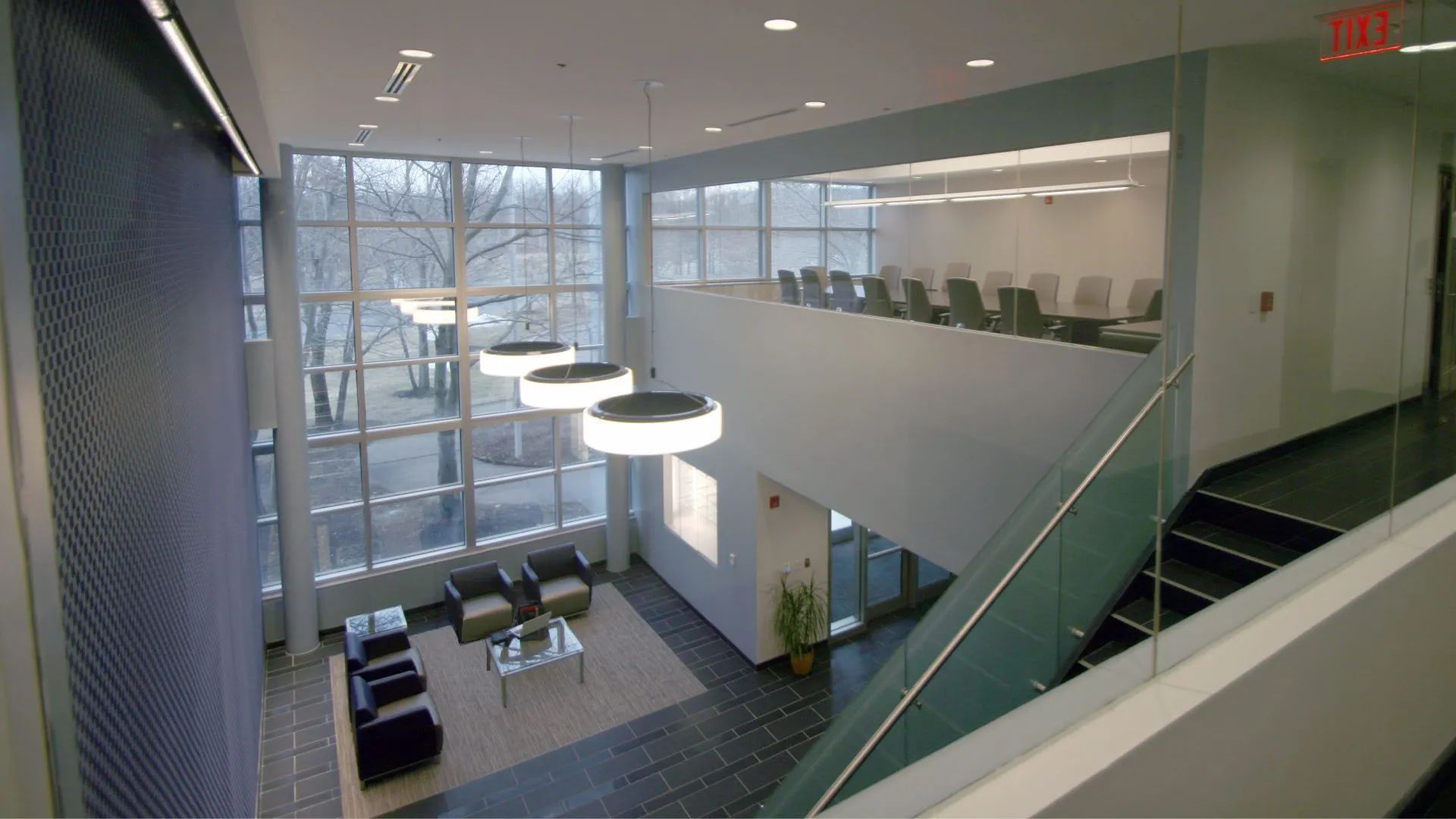 Modern office lobby with glass walls, stairs, lounge seating, and pendant lights from an upper walkway