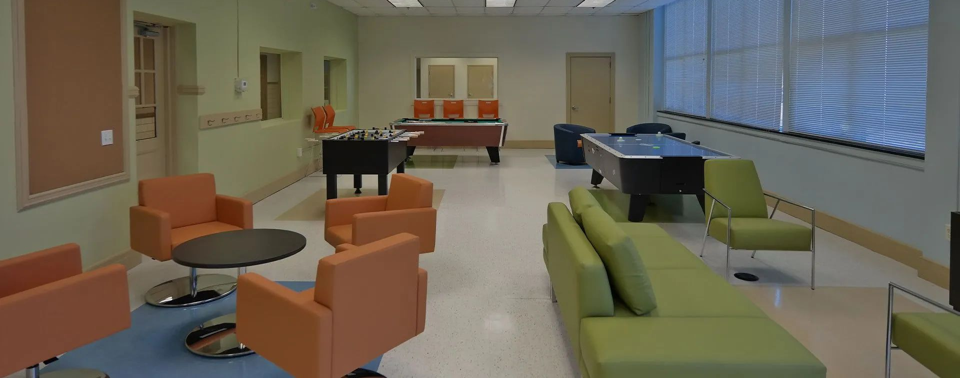 Modern lounge with orange and green chairs, tables, couches, and large windows in a bright room