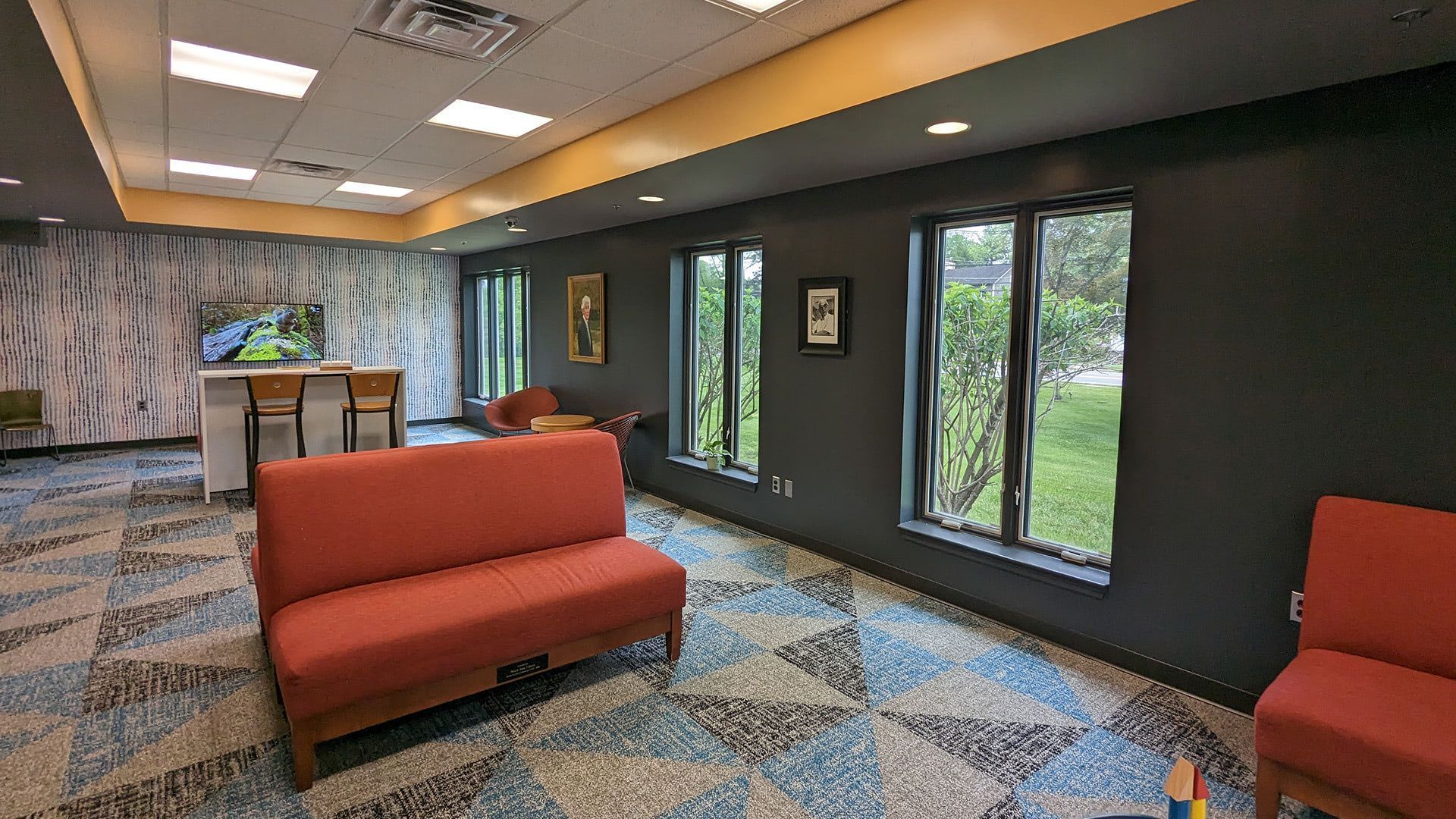 Renovated interior showing the bright, modern reading area with orange seating, patterned blue carpet, large windows, and the counter area. Rocky Hill Library — Rocky Hill, NJ