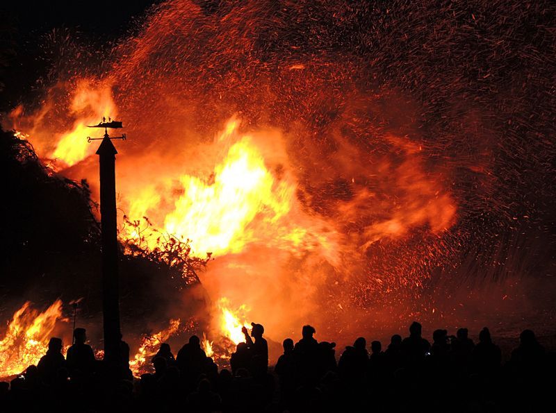 A crowd of people stands in silhouette before a massive, bright orange bonfire with sparks flying into the night sky.