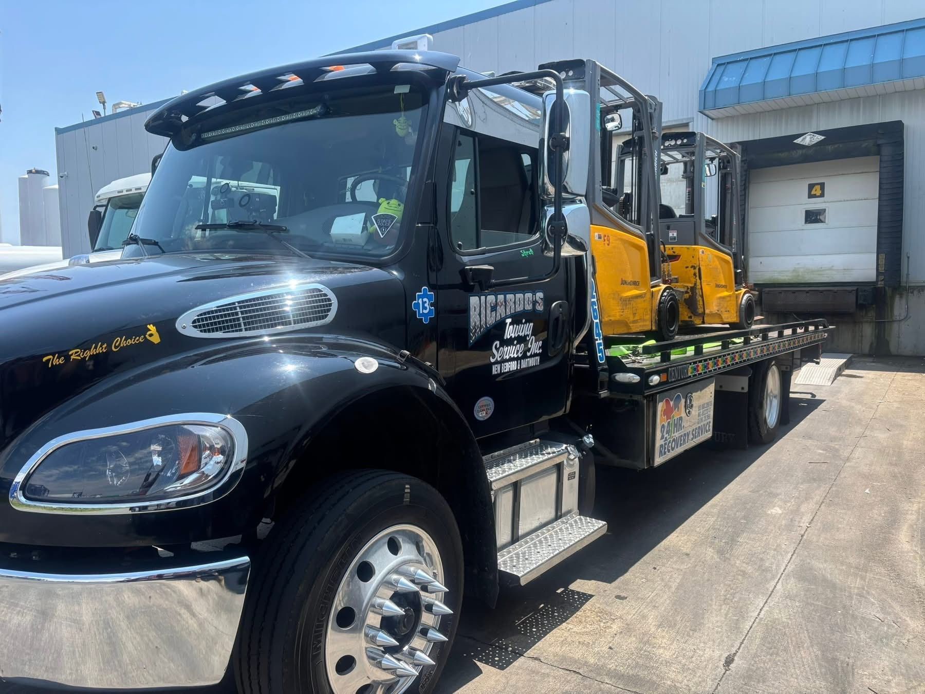 Black flatbed tow truck loaded with yellow equipment parked at a loading dock