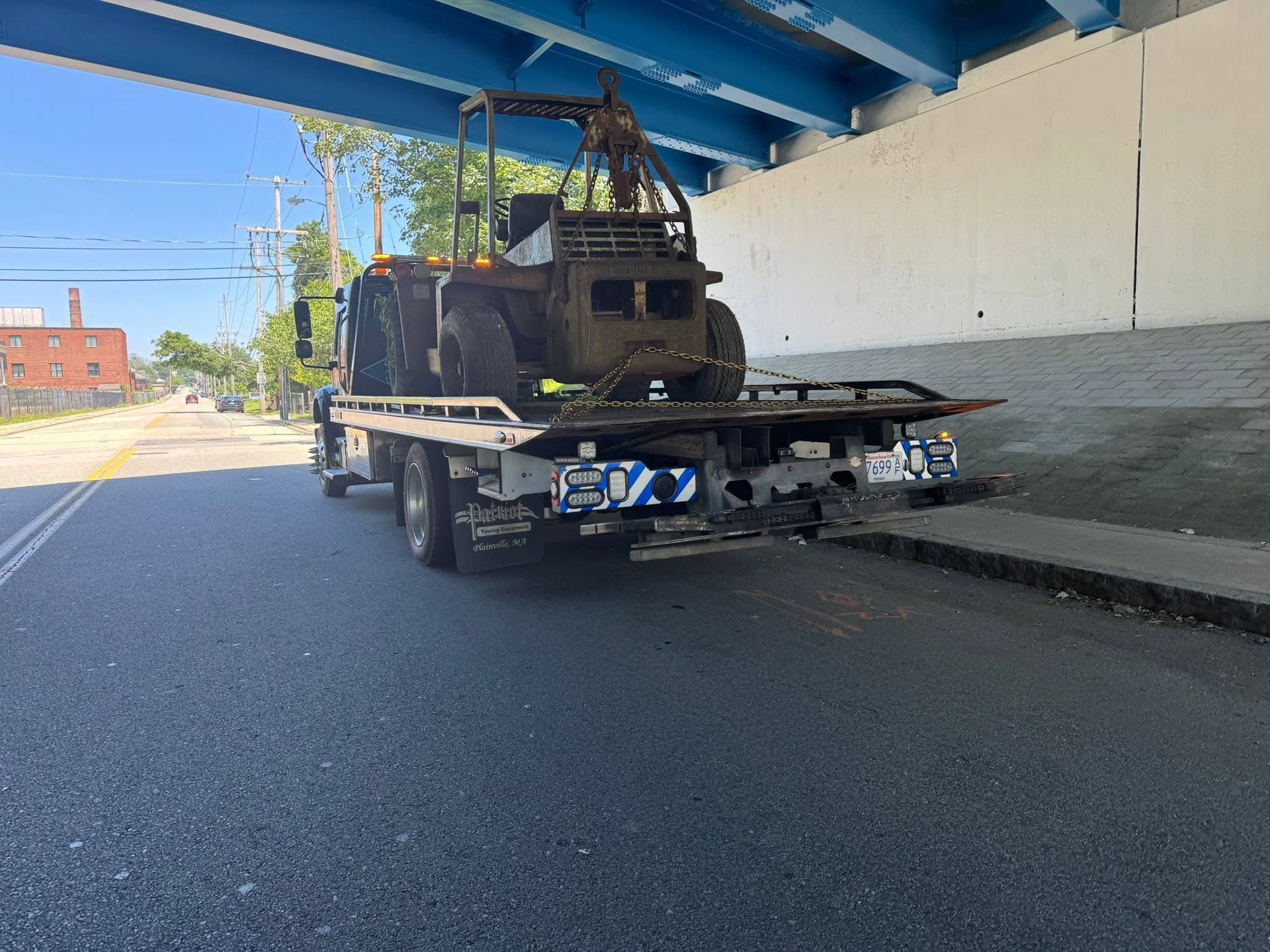 Flatbed tow truck carrying a burned-out vehicle under a blue overpass on a city street