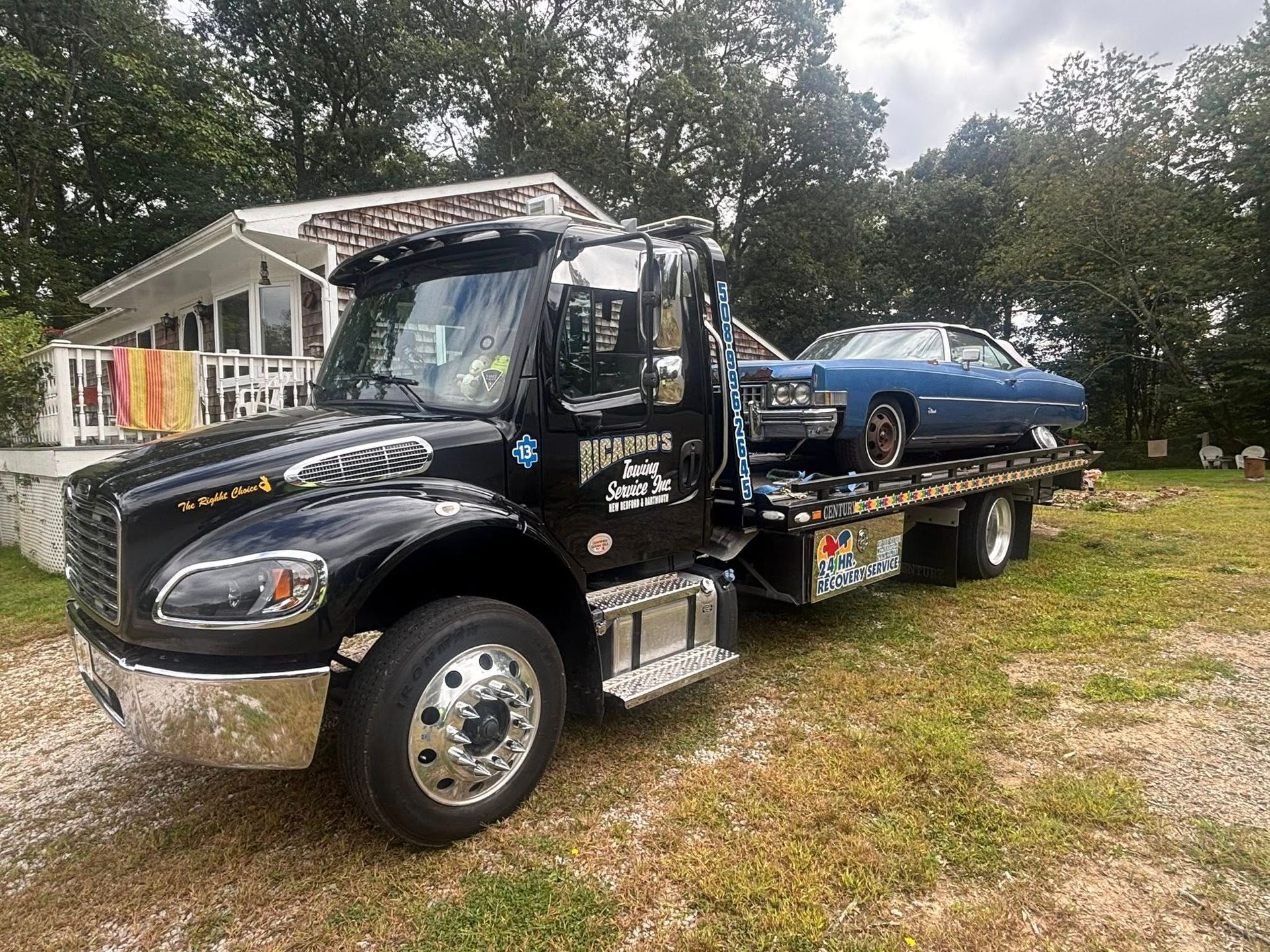 Black tow truck hauling a blue car beside a house on a grassy lot