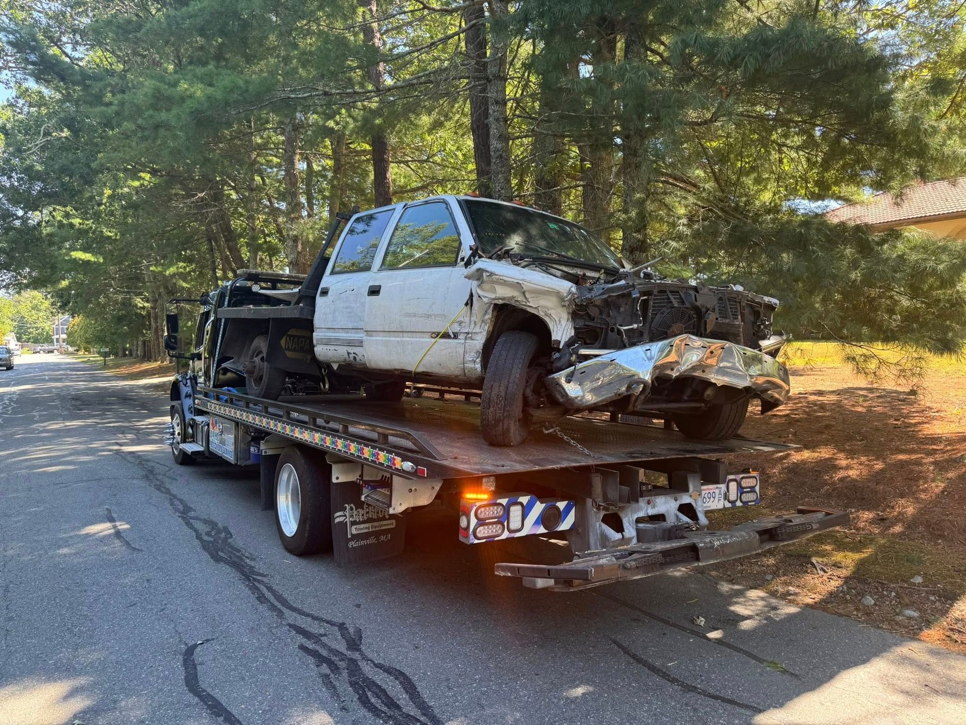 White pickup truck with front-end damage on a flatbed tow truck beside a wooded road