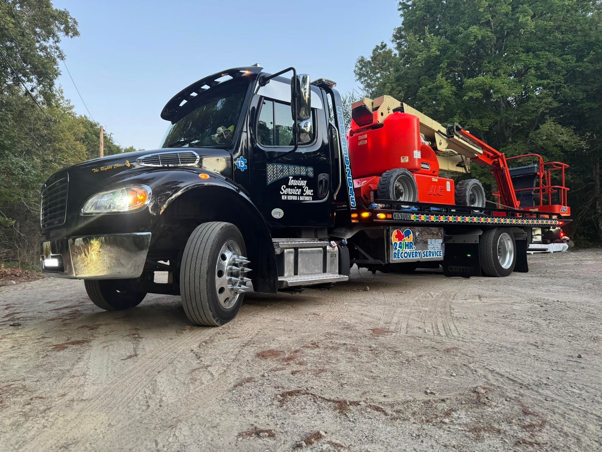 Black tow truck carrying red construction equipment on a gravel road under trees at dusk