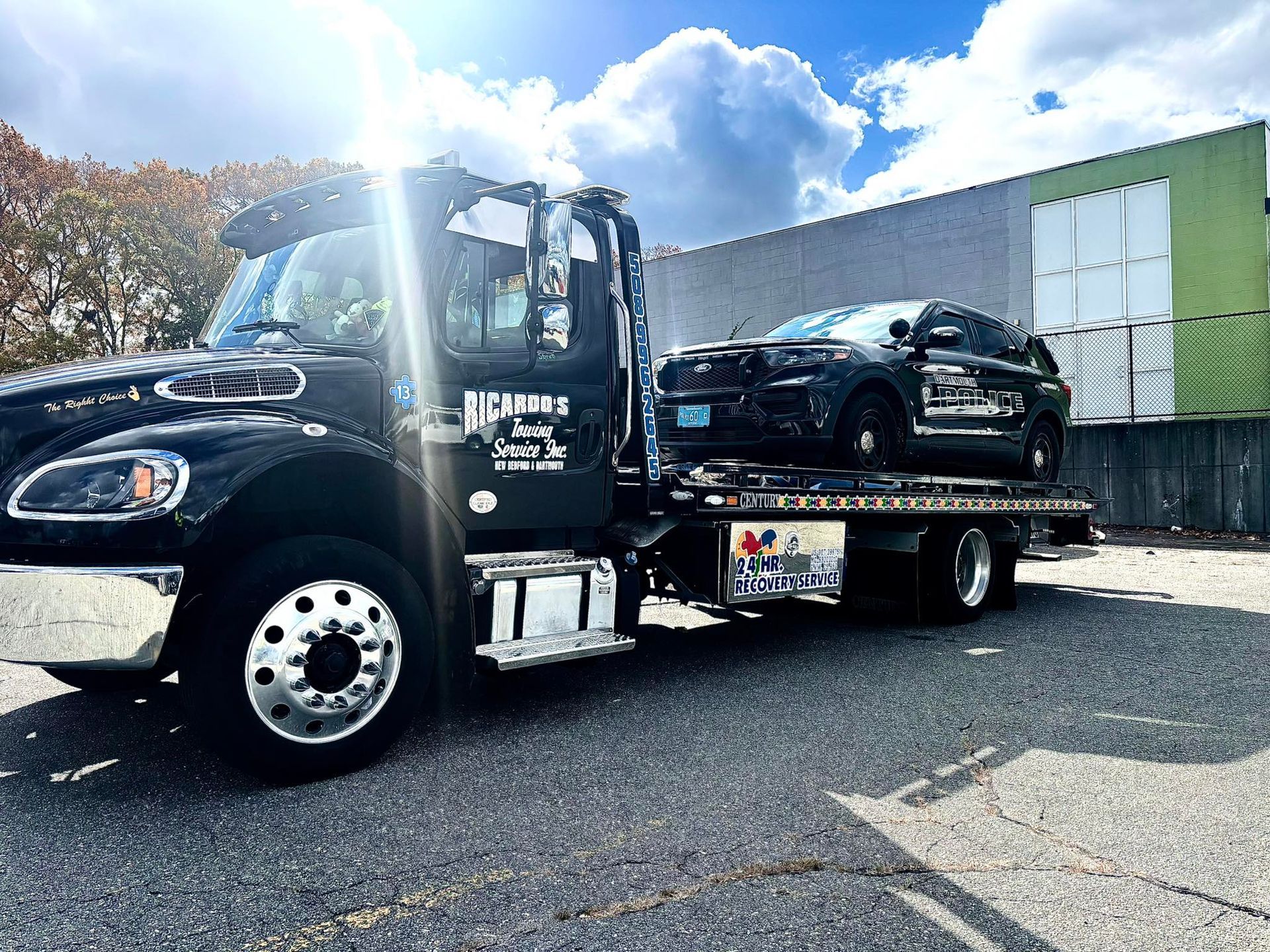 Black tow truck hauling a black pickup on a flatbed in a sunny parking lot