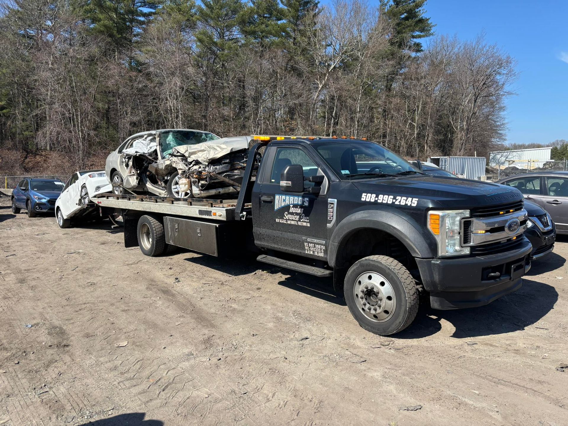 Tow truck hauling a crushed white car on a dirt lot beside a snowy road, with trees in the background.