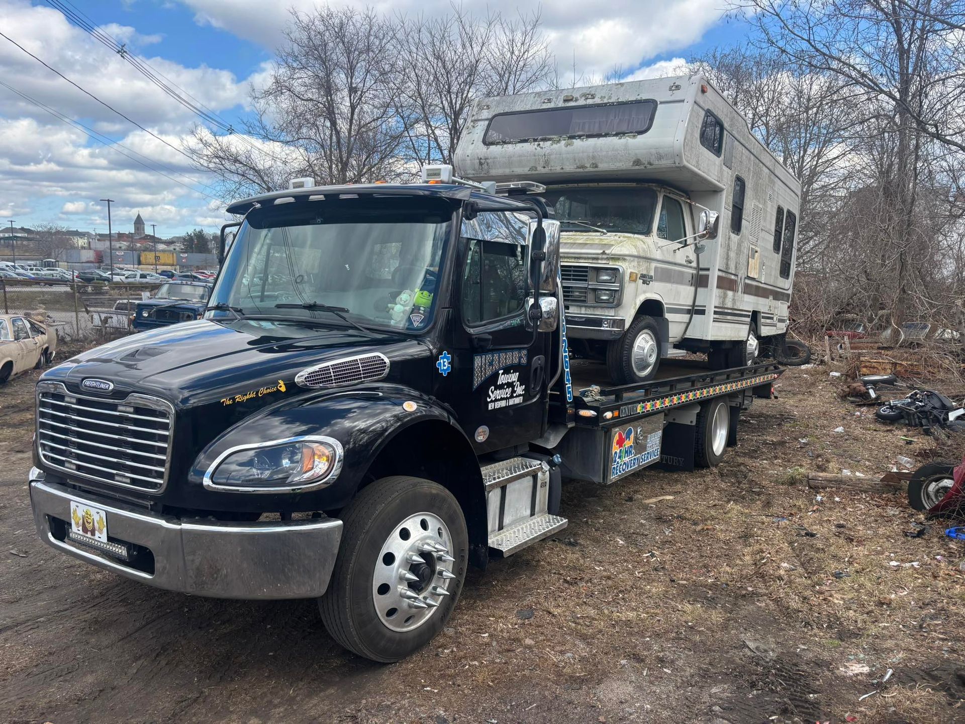 Black tow truck carrying a white RV on a flatbed in a rocky outdoor lot under a cloudy sky