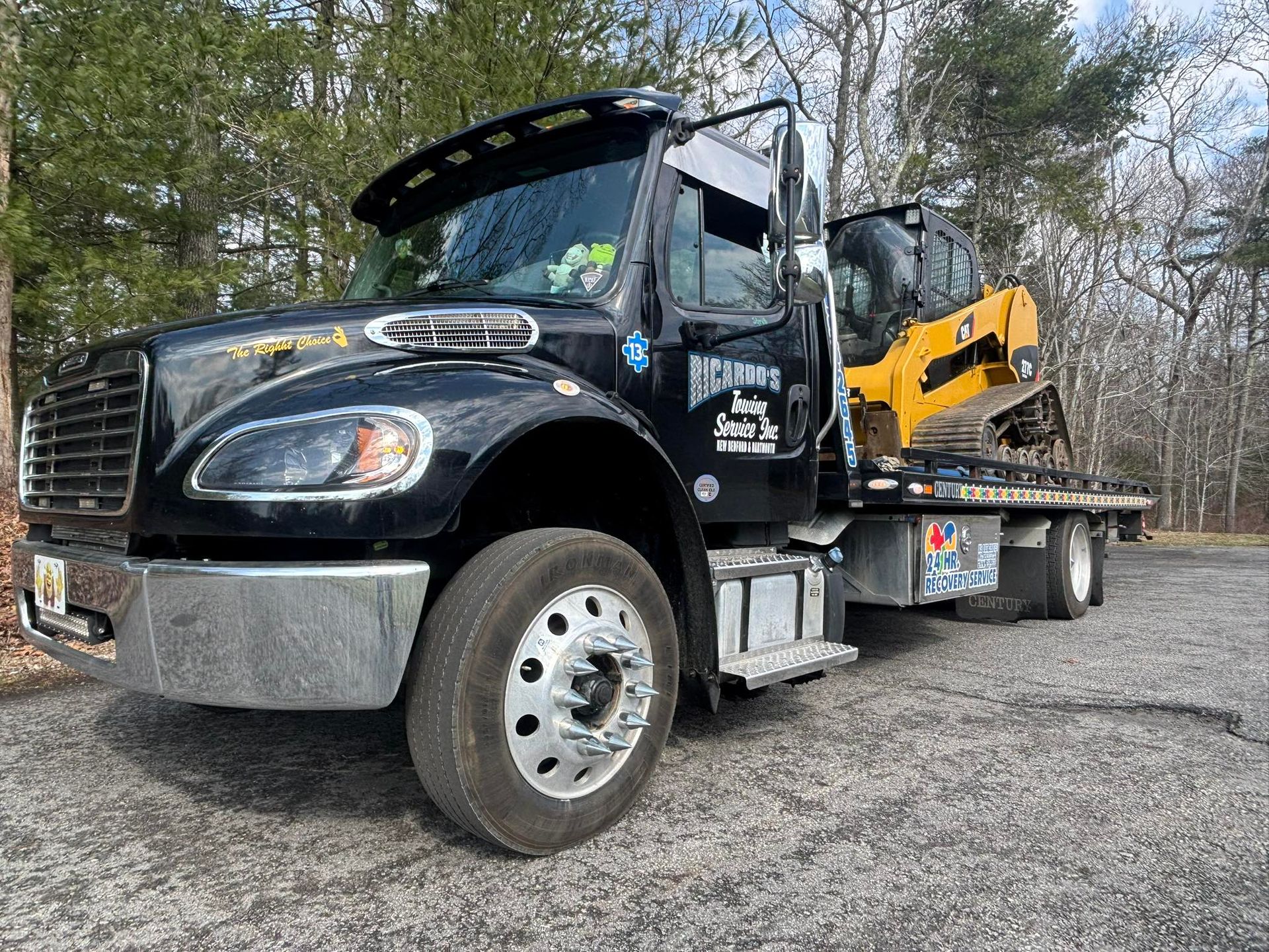 Black tow truck hauling a yellow skid steer on a flatbed in a gravel lot