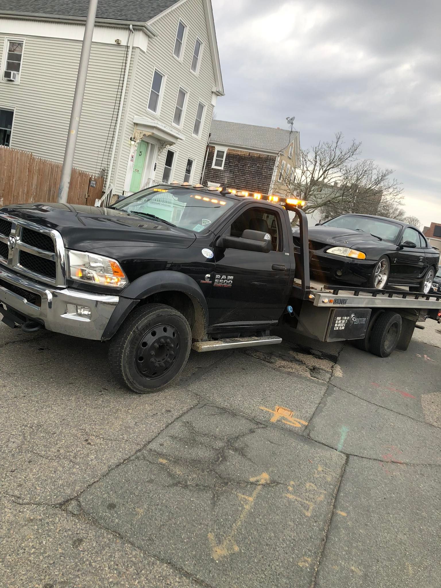 Black tow truck carrying a car on a flatbed parked on a city street near houses at dusk