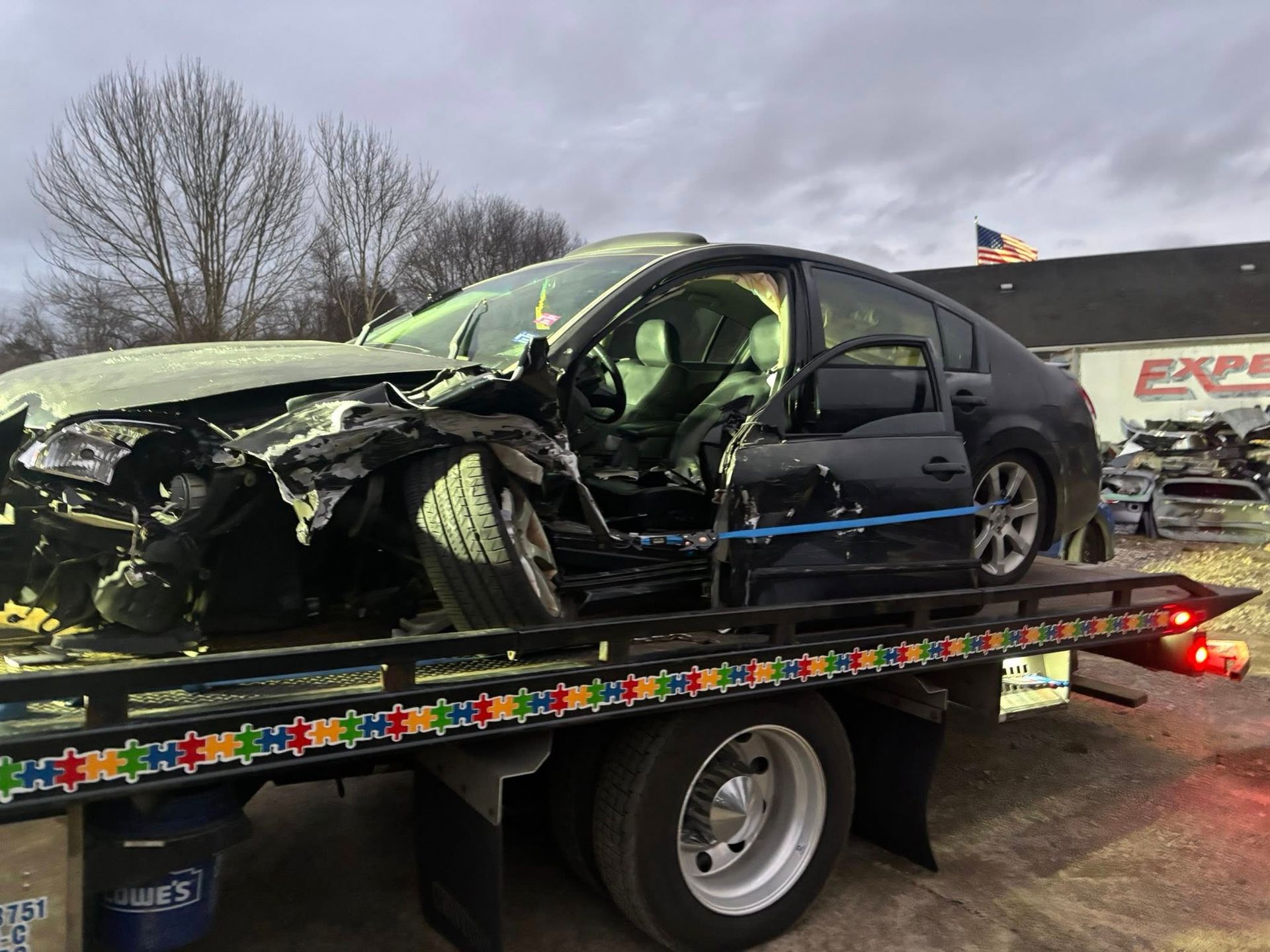 Wrecked black sedan on a tow truck, front end badly damaged, with a damaged truck in the background.