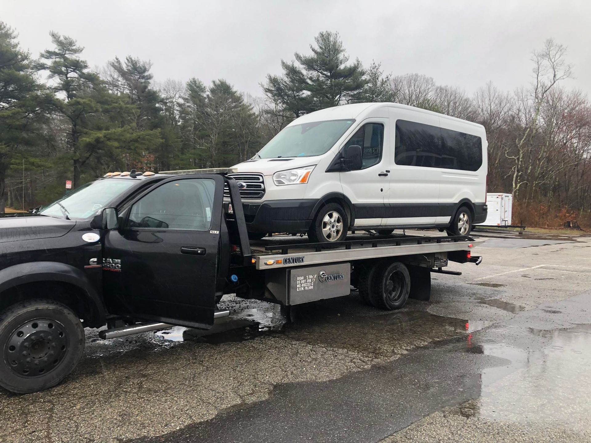 Black tow truck carrying a white van on a wet roadside in a wooded area