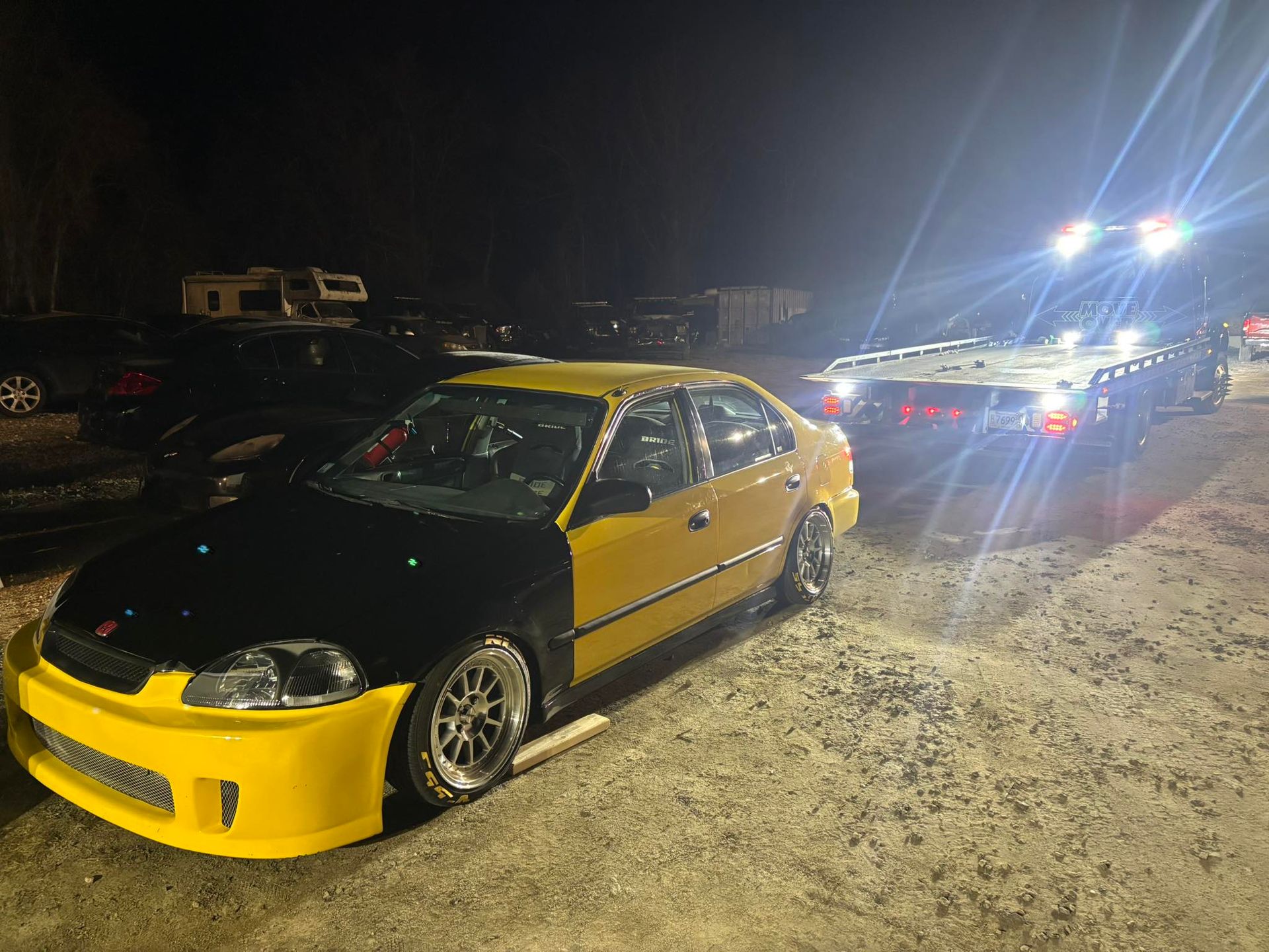 Yellow-and-black modified car at night on a snowy roadside, with headlights from vehicles behind it
