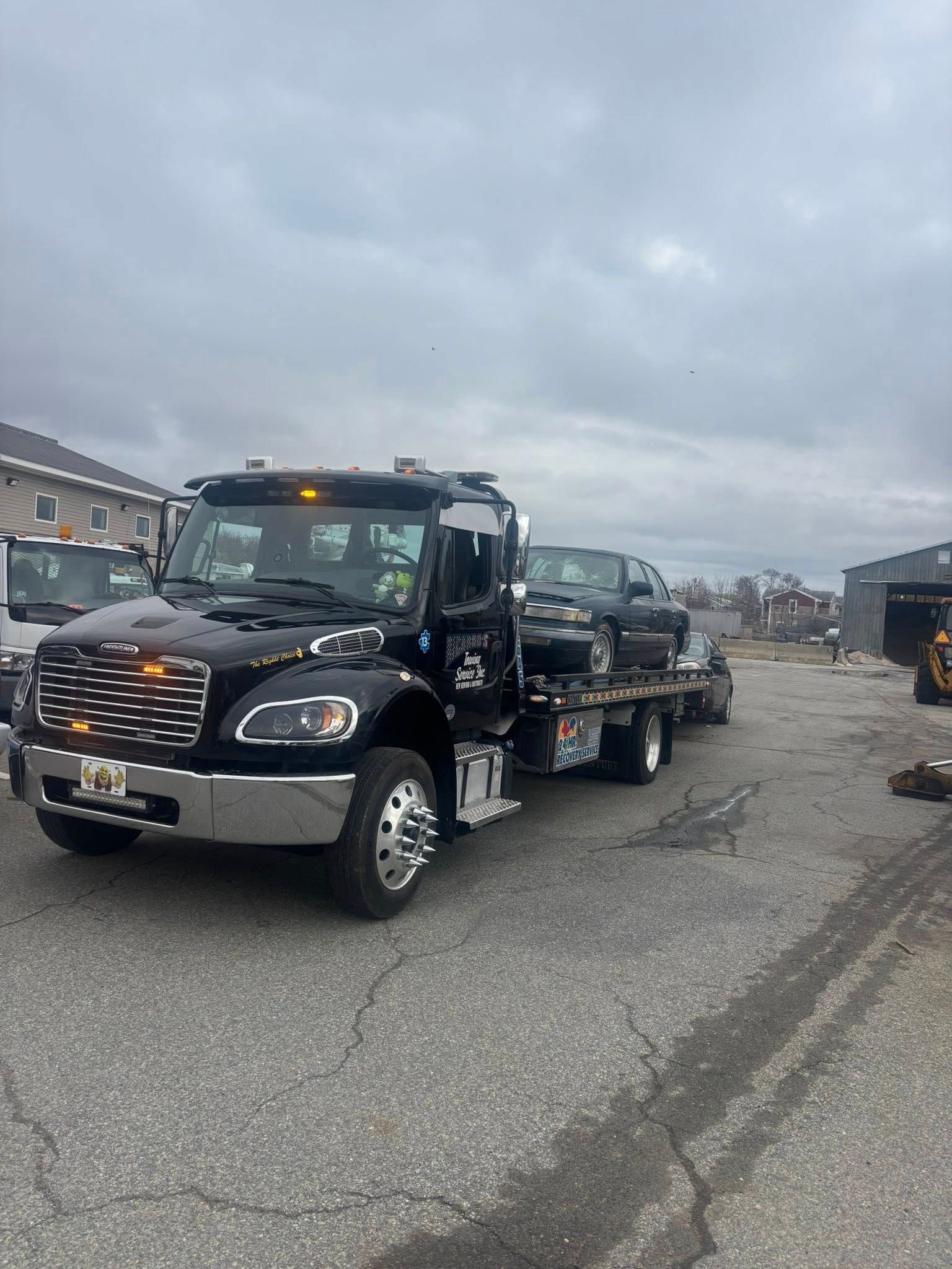 Black tow truck hauling a damaged car on a cloudy roadside lot