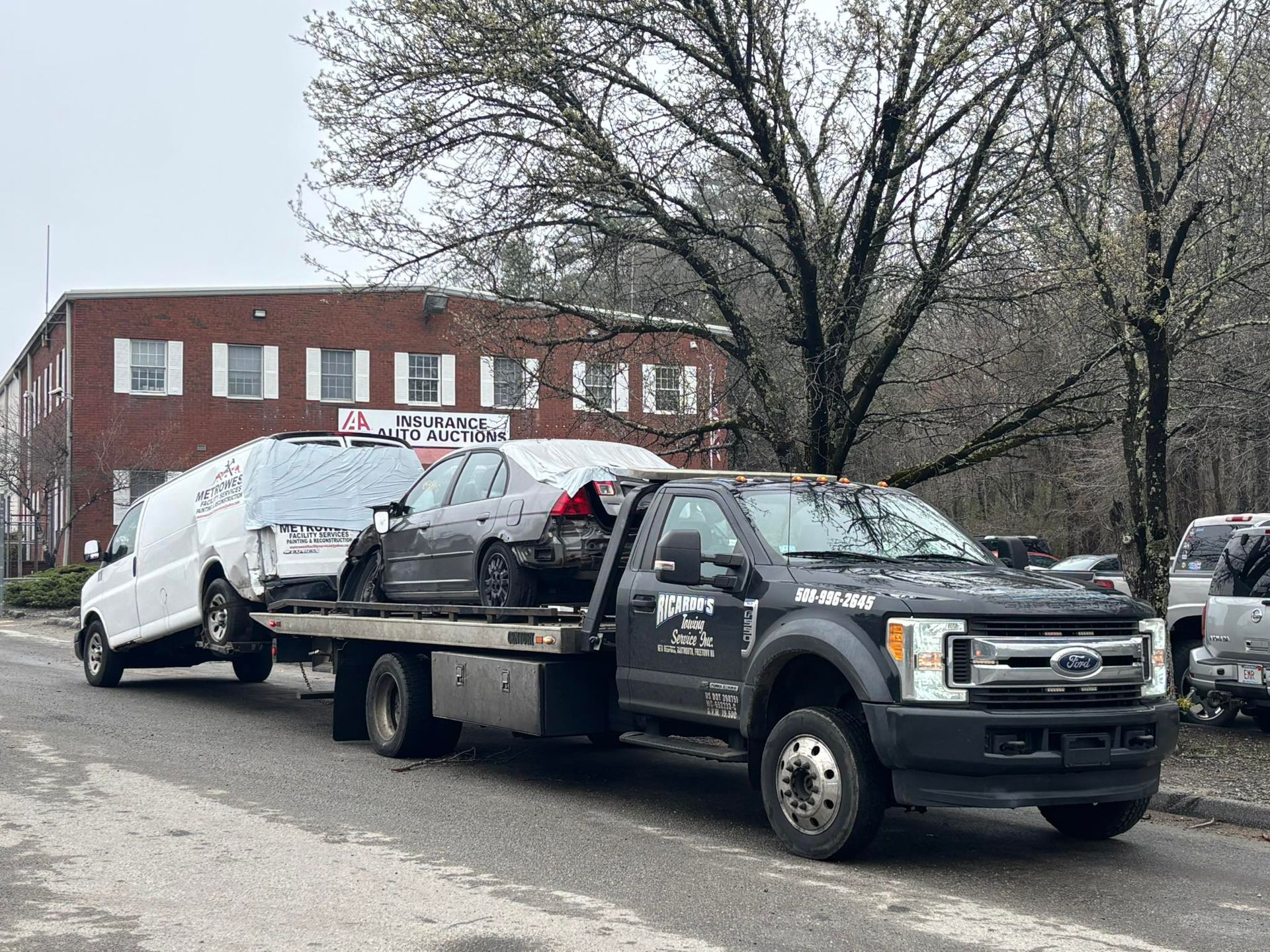 Tow truck hauling a white car on a flatbed in front of a brick building and bare trees