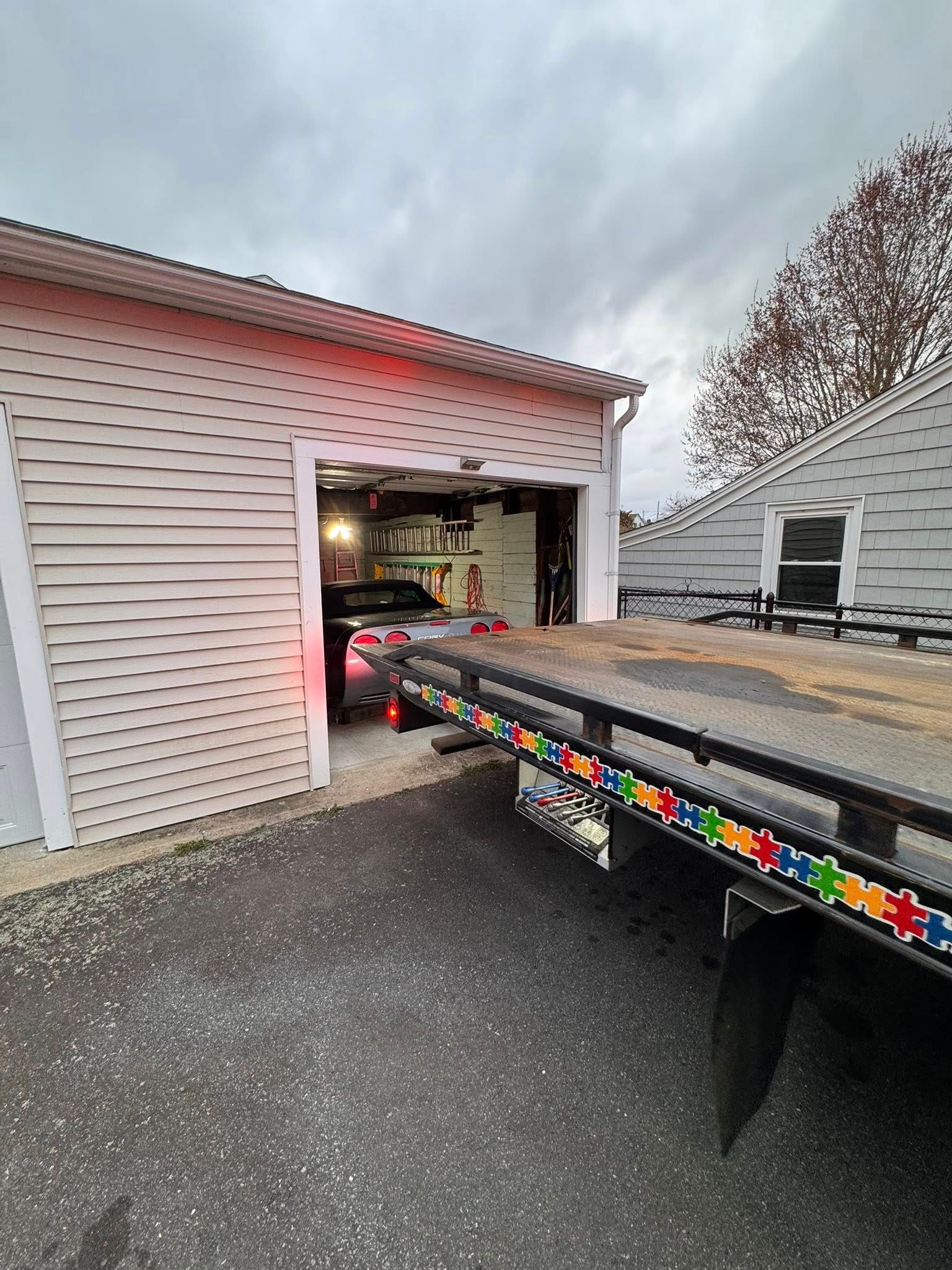 Tow truck backing into an open garage beside a white house on a cloudy day