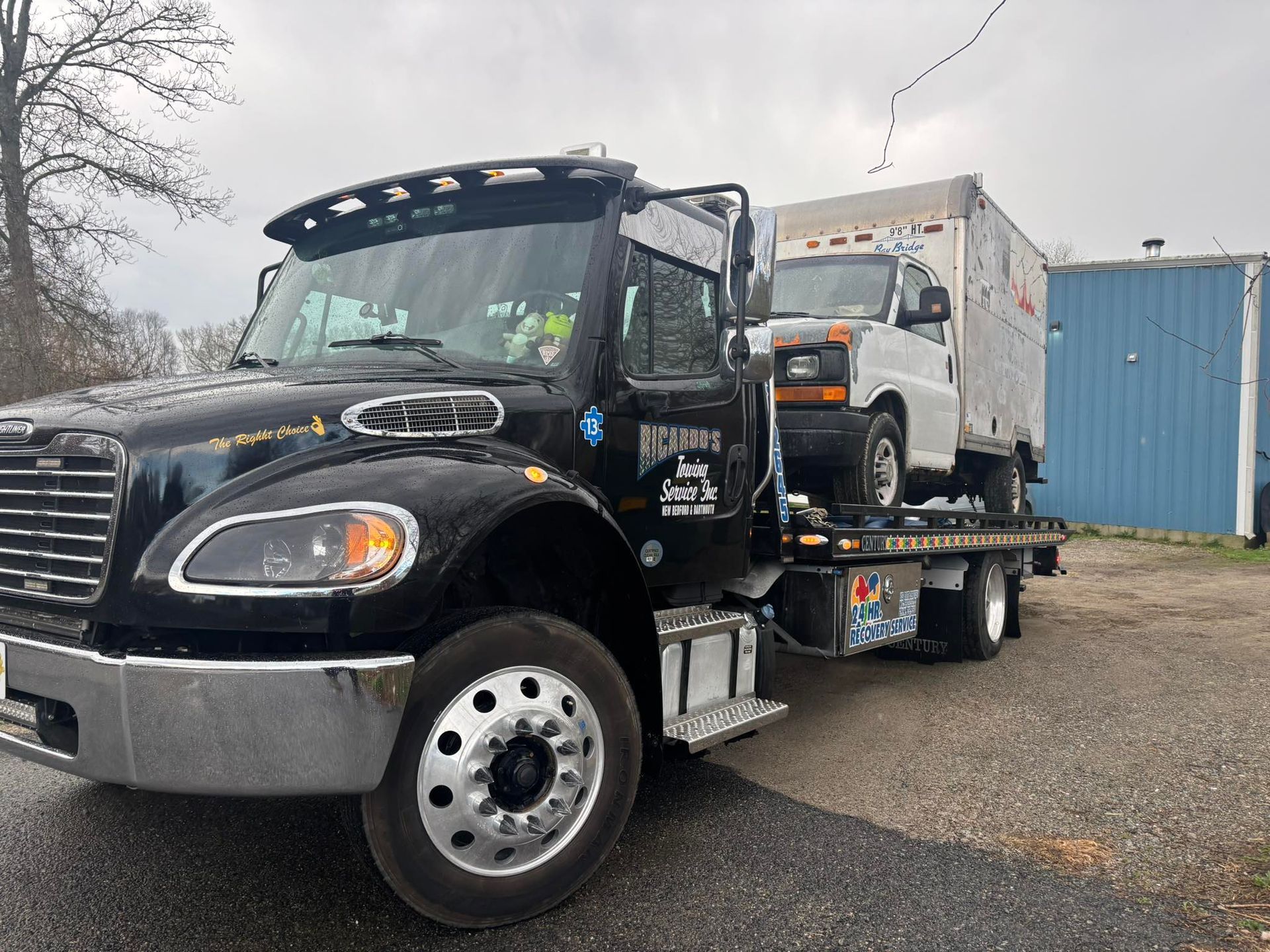 Black flatbed tow truck carrying a white van in a gravel lot on an overcast day
