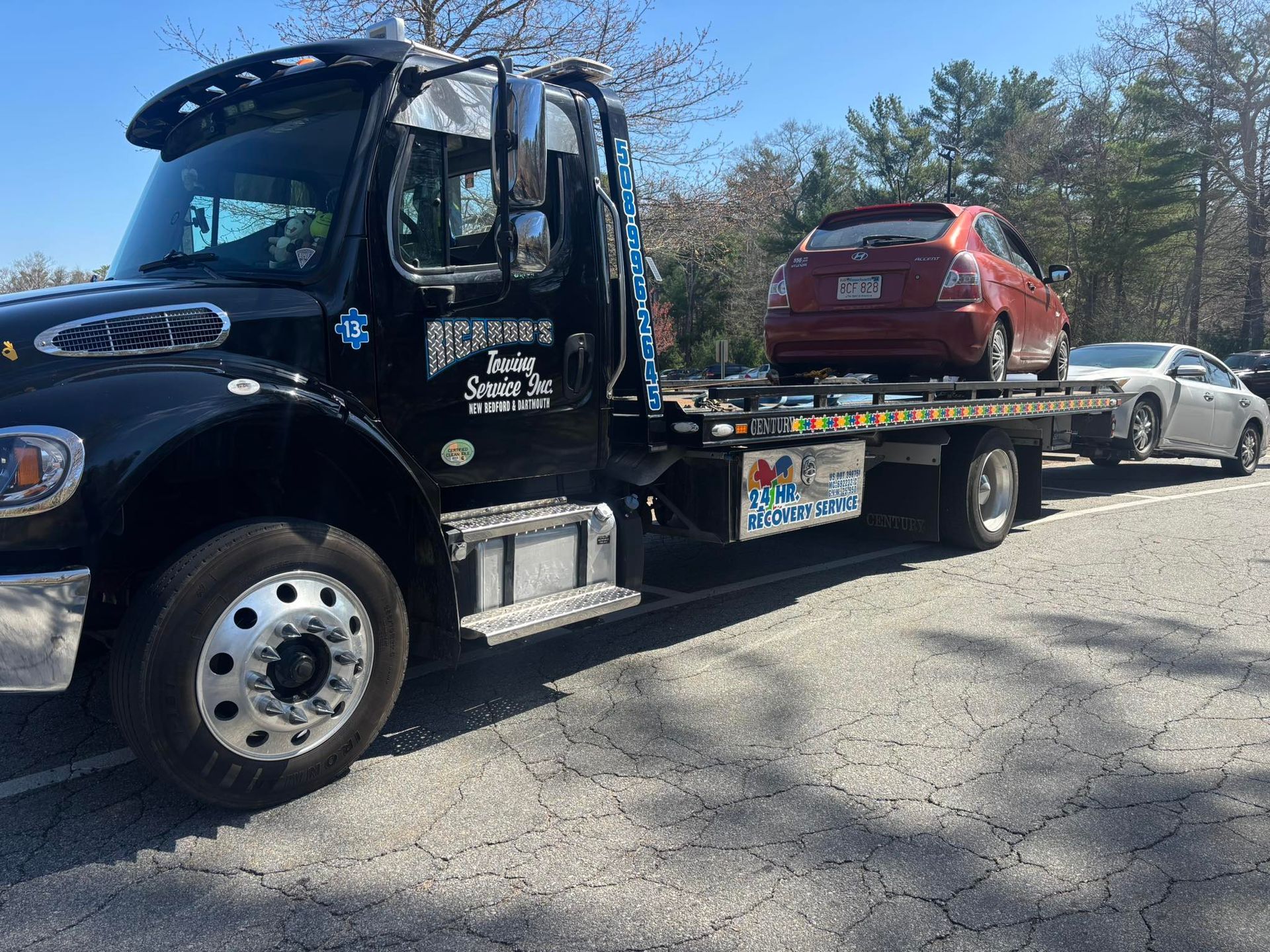 Black tow truck carrying a red SUV on a gravel roadside near trees