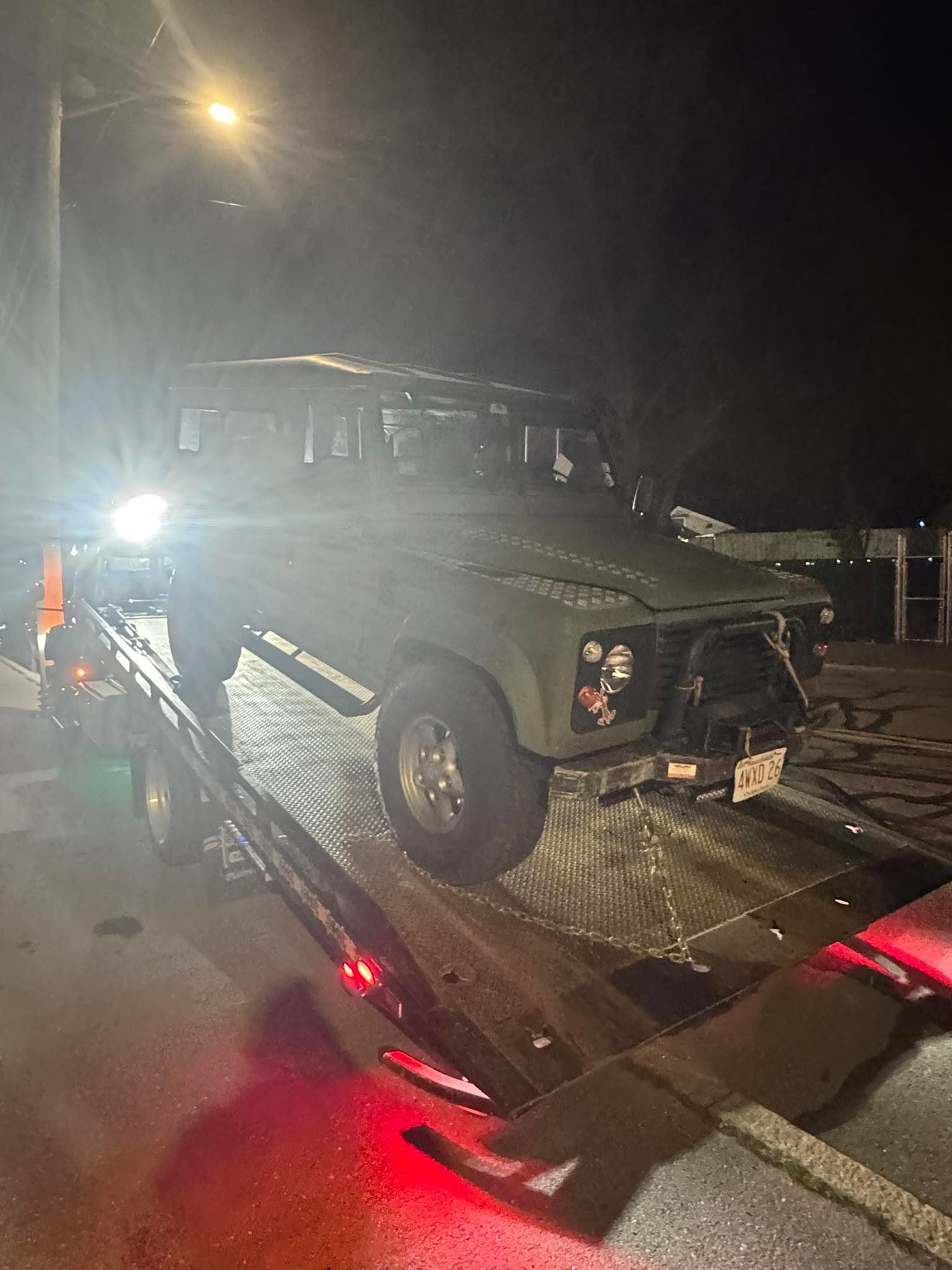 Dark green off-road vehicle on a tow truck at night, with headlights and red reflections on wet pavement