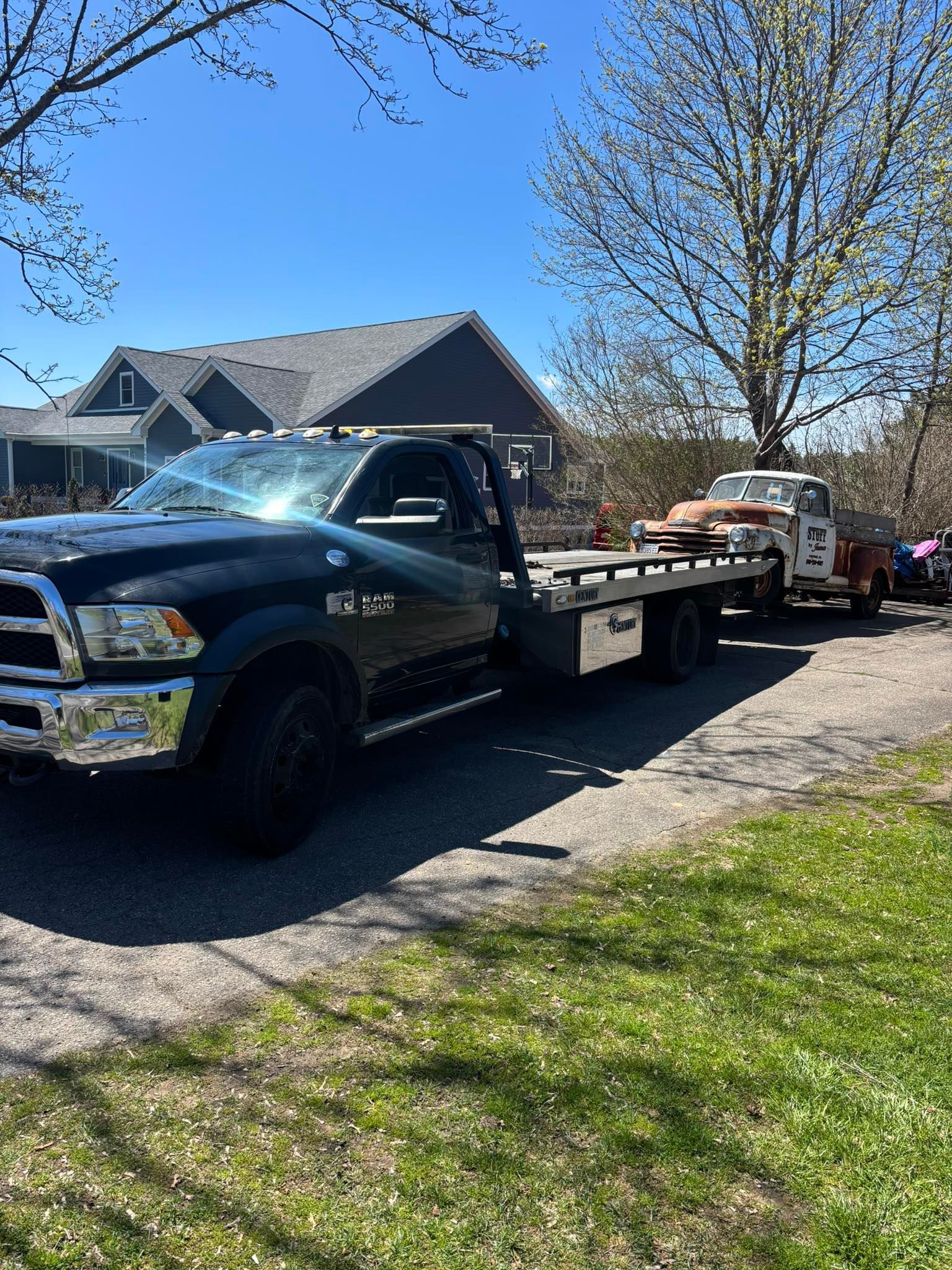 Black flatbed tow truck parked on a residential street beside a house under a blue sky.