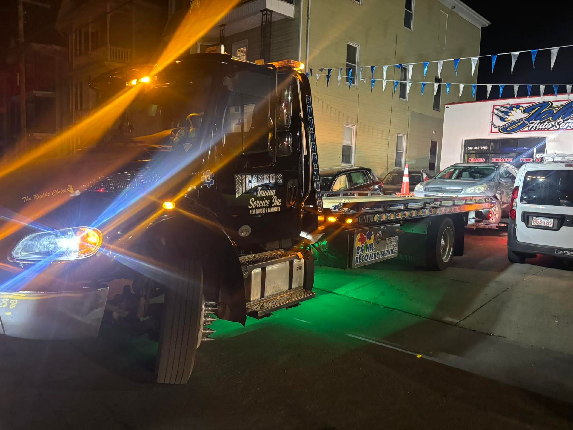 Tow truck at night with flashing lights towing a car on a city street and lit building in background