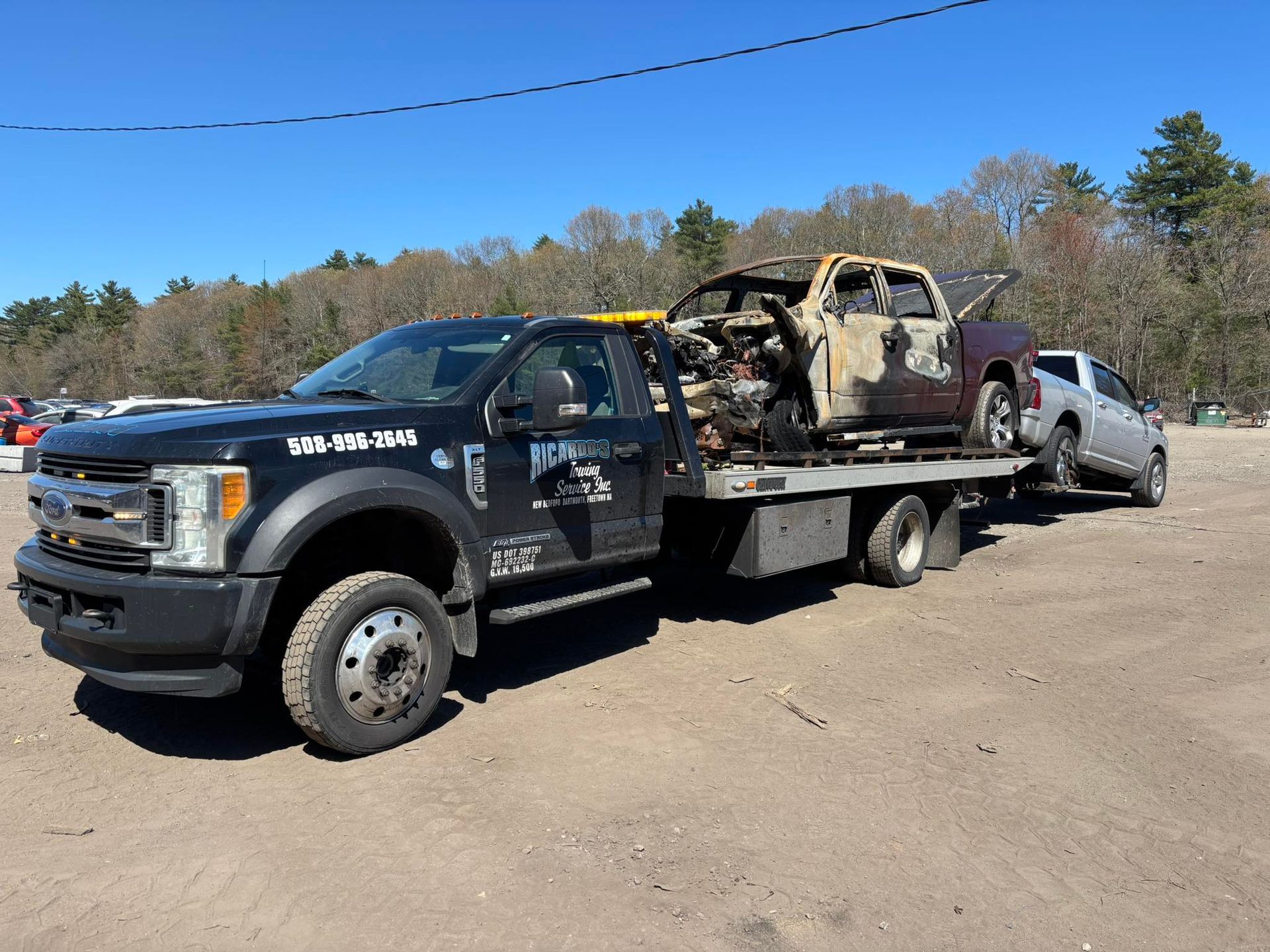 Tow truck hauling a burned-out pickup and a damaged silver car on a dirt lot.