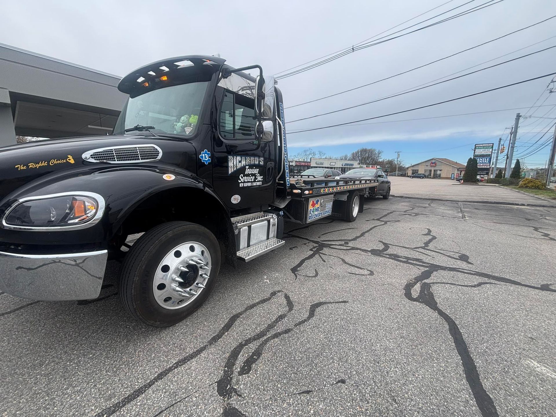 Black flatbed tow truck parked on cracked pavement under cloudy sky.