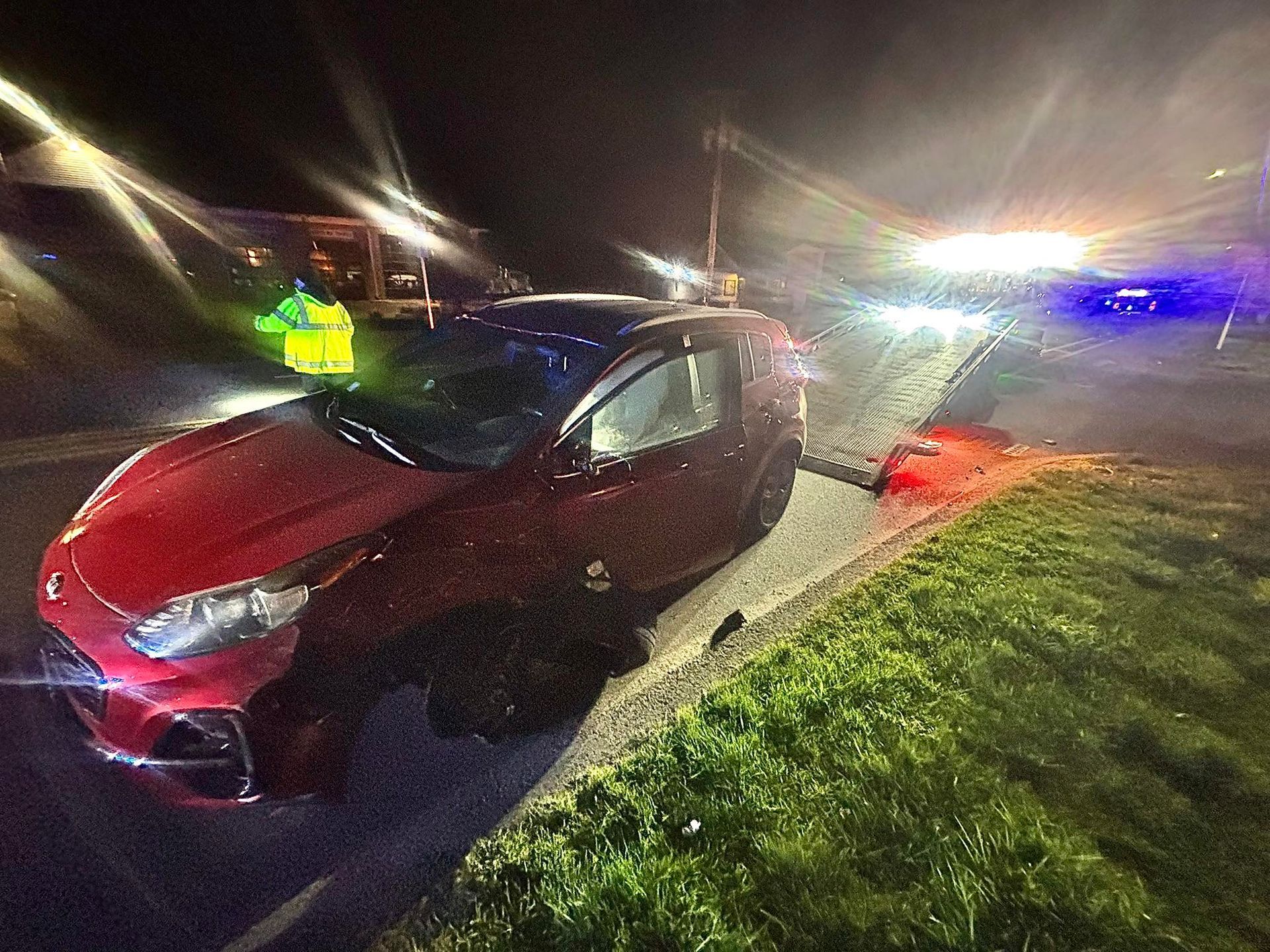 Red car crashed on a roadside at night, with emergency lights and a responder nearby.