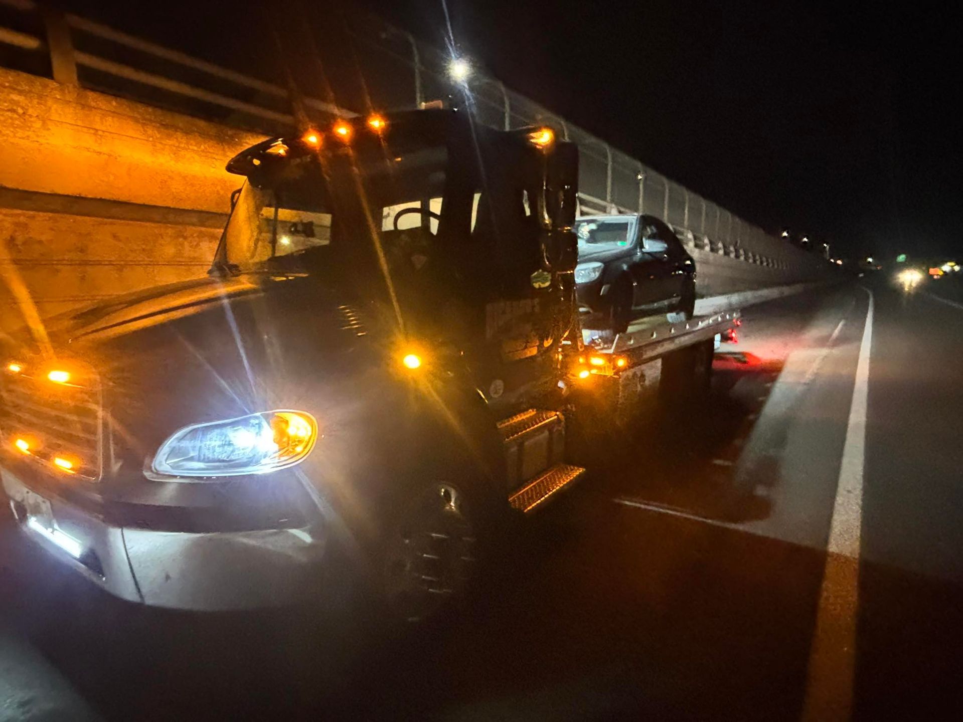 Tow truck hauling a silver car on a highway at night, amber hazard lights glowing