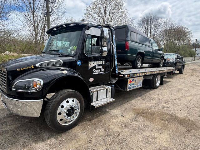 Black tow truck carrying a dark SUV on a flatbed in a gravel lot under cloudy skies