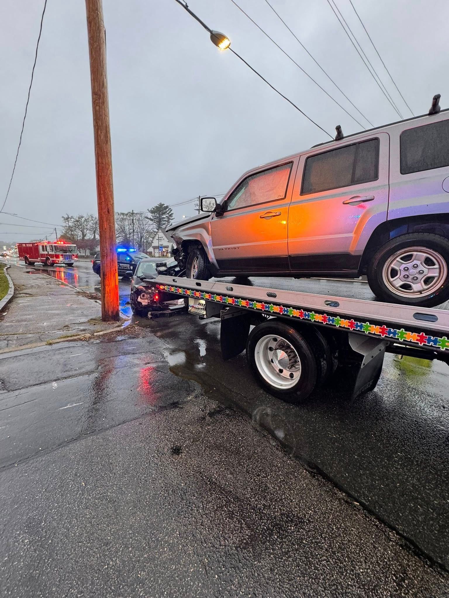 Tow truck lifting a silver SUV in a rainy street beside a utility pole and police lights