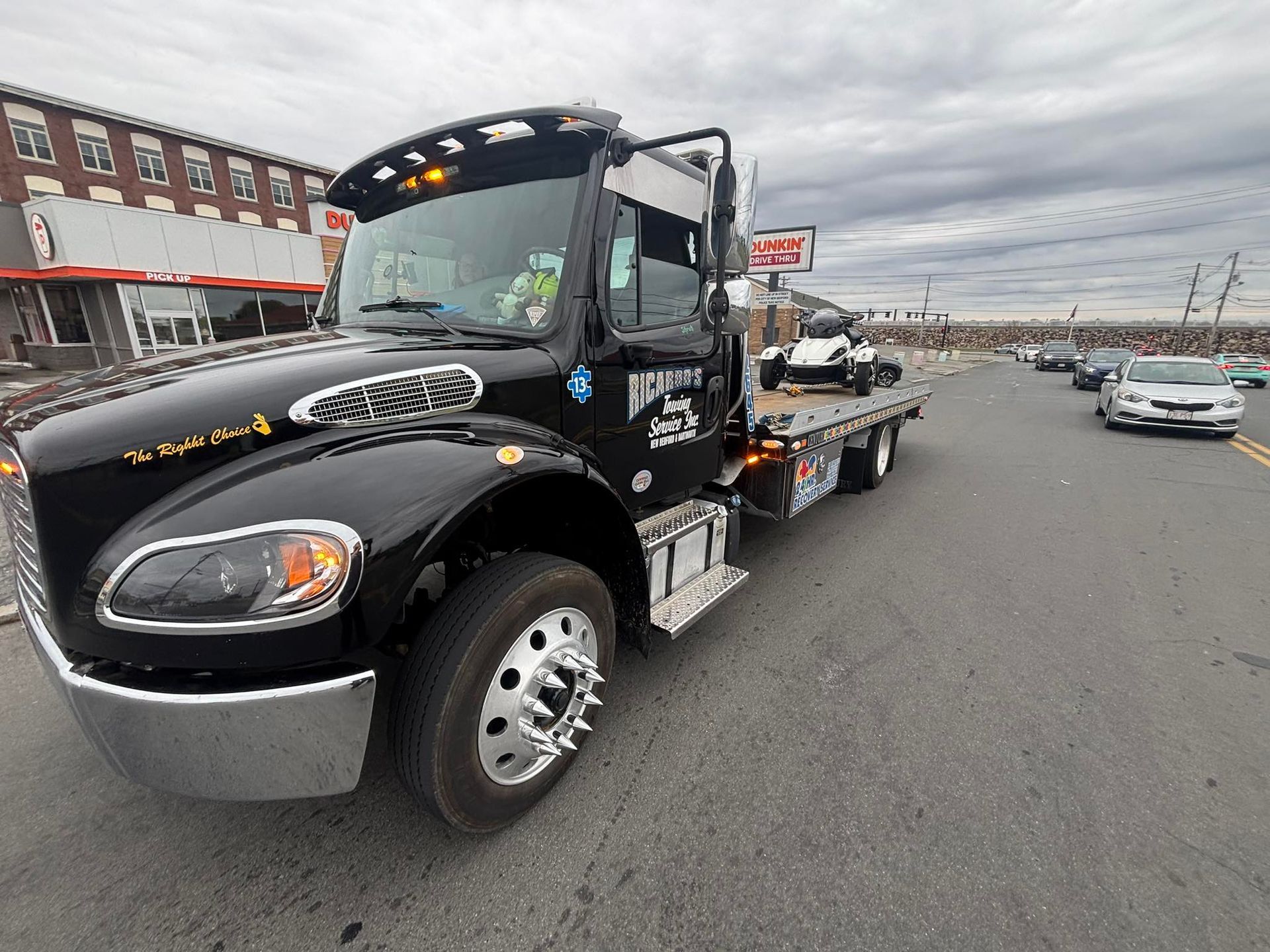 Black flatbed tow truck parked on a street beside shops and traffic under a cloudy sky