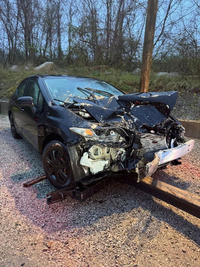 Severely crashed black car with crushed front end on a gravel roadside near trees