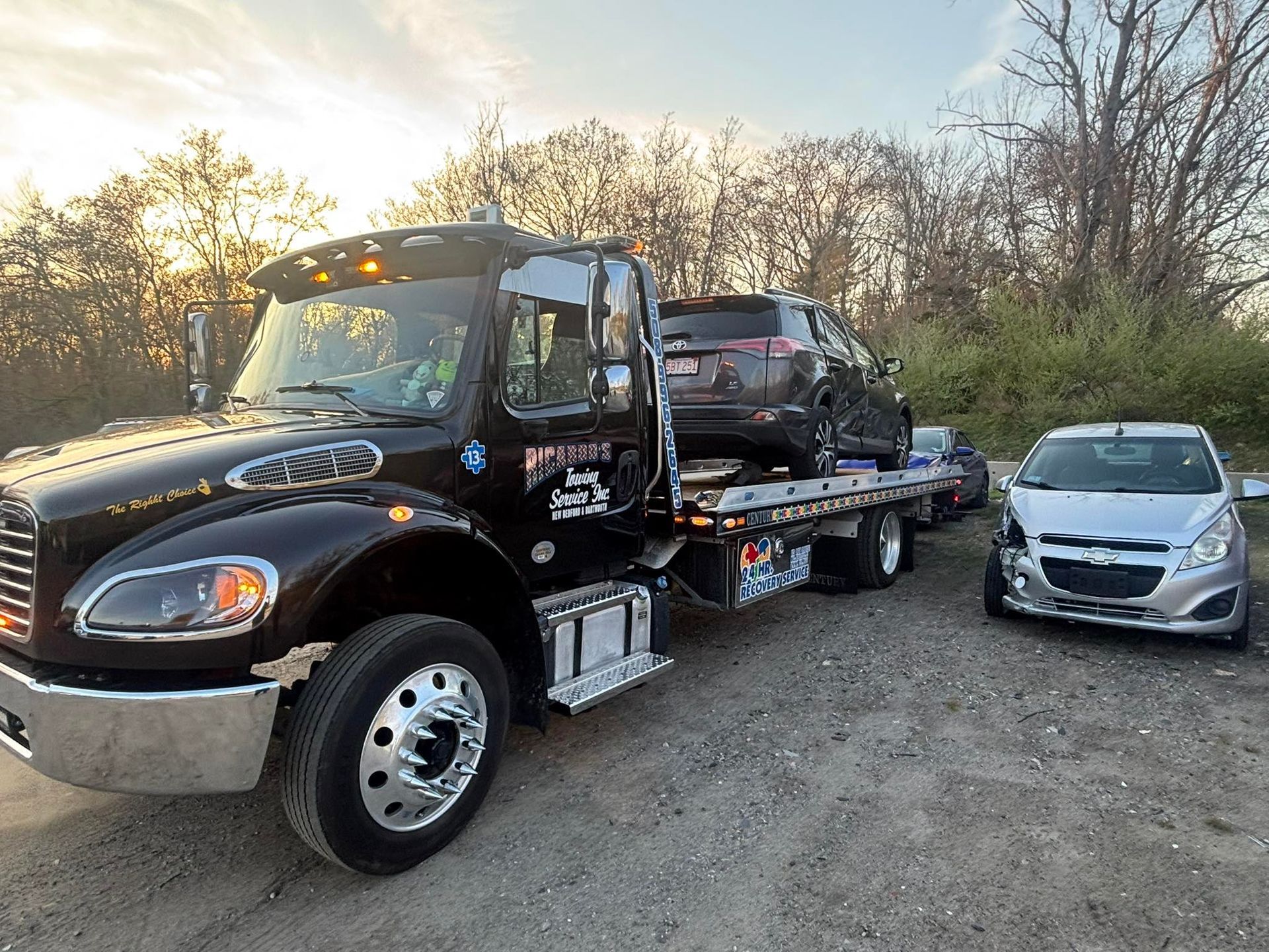 Tow truck hauling a black car beside a silver compact car on a gravel roadside at sunset