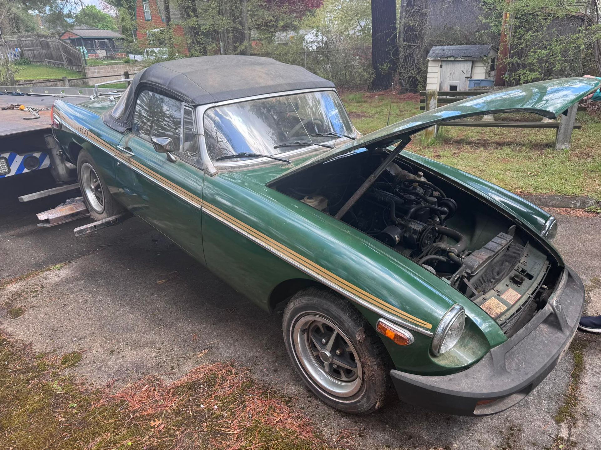 Green classic convertible with hood open, parked in a driveway on a rainy day
