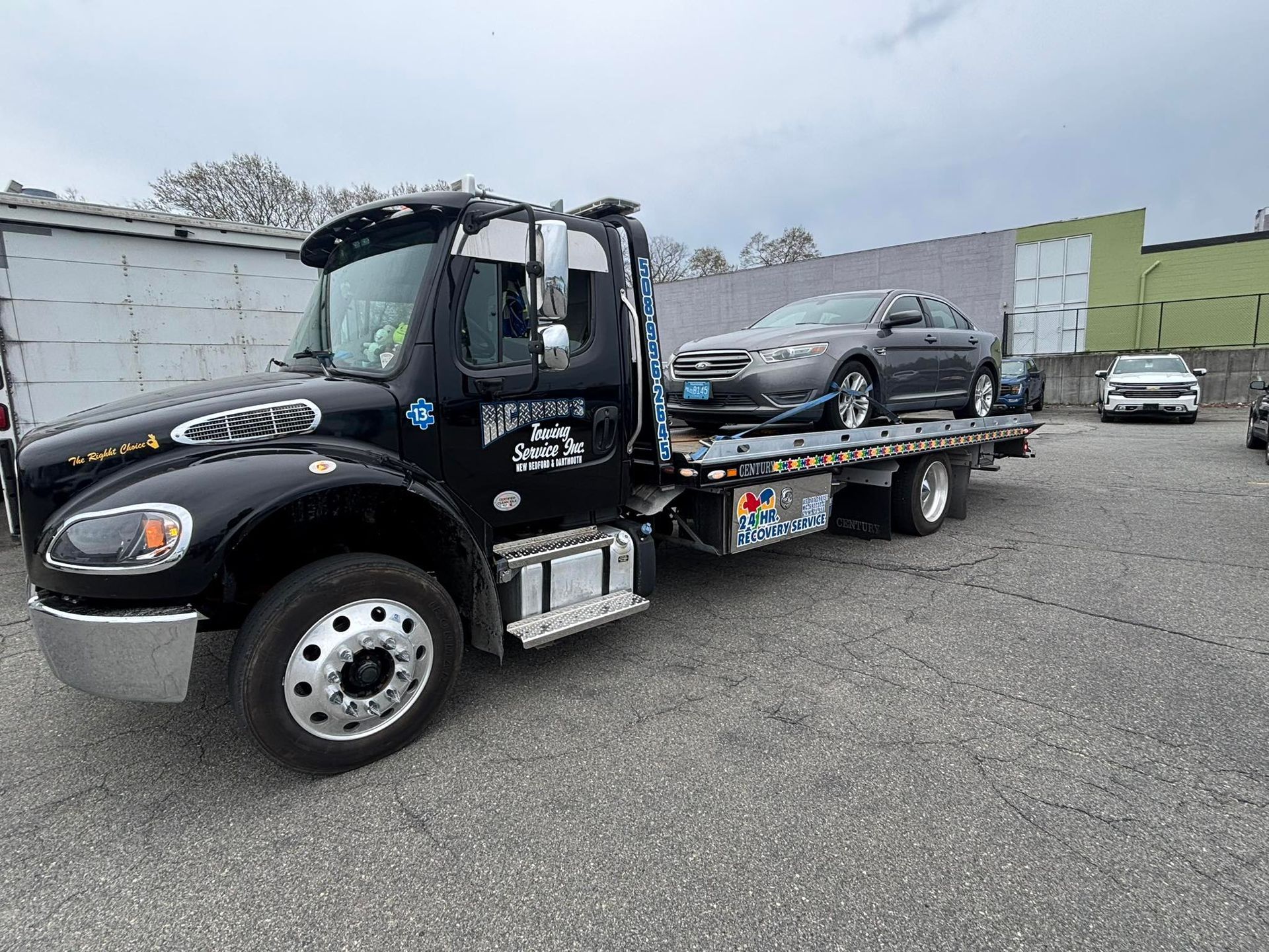 Black tow truck carrying a silver car in a gravel lot under an overcast sky