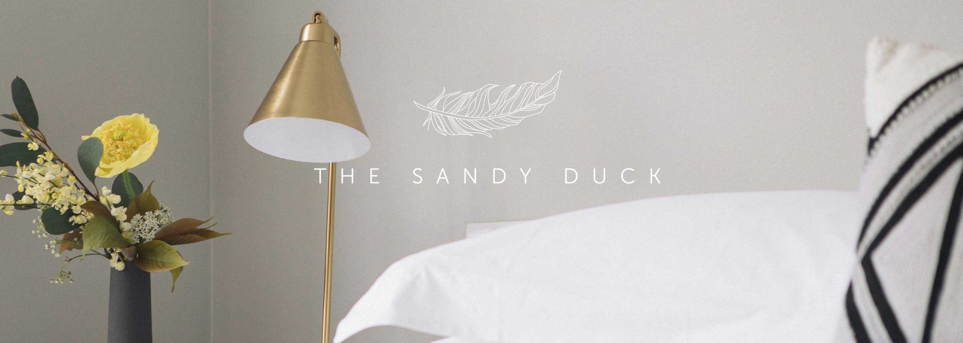 Gold lamp beside a white bed in a soft, neutral bedroom with flowers on a side table