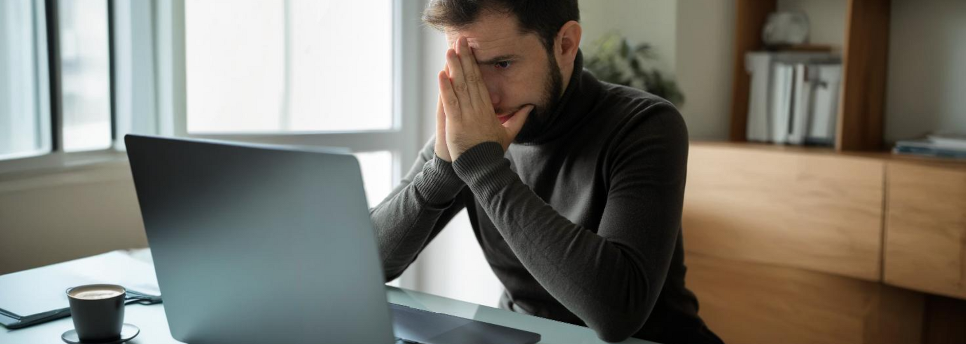 Man sitting at a desk, leaning on his hand and looking at a laptop, appearing stressed in a home office.