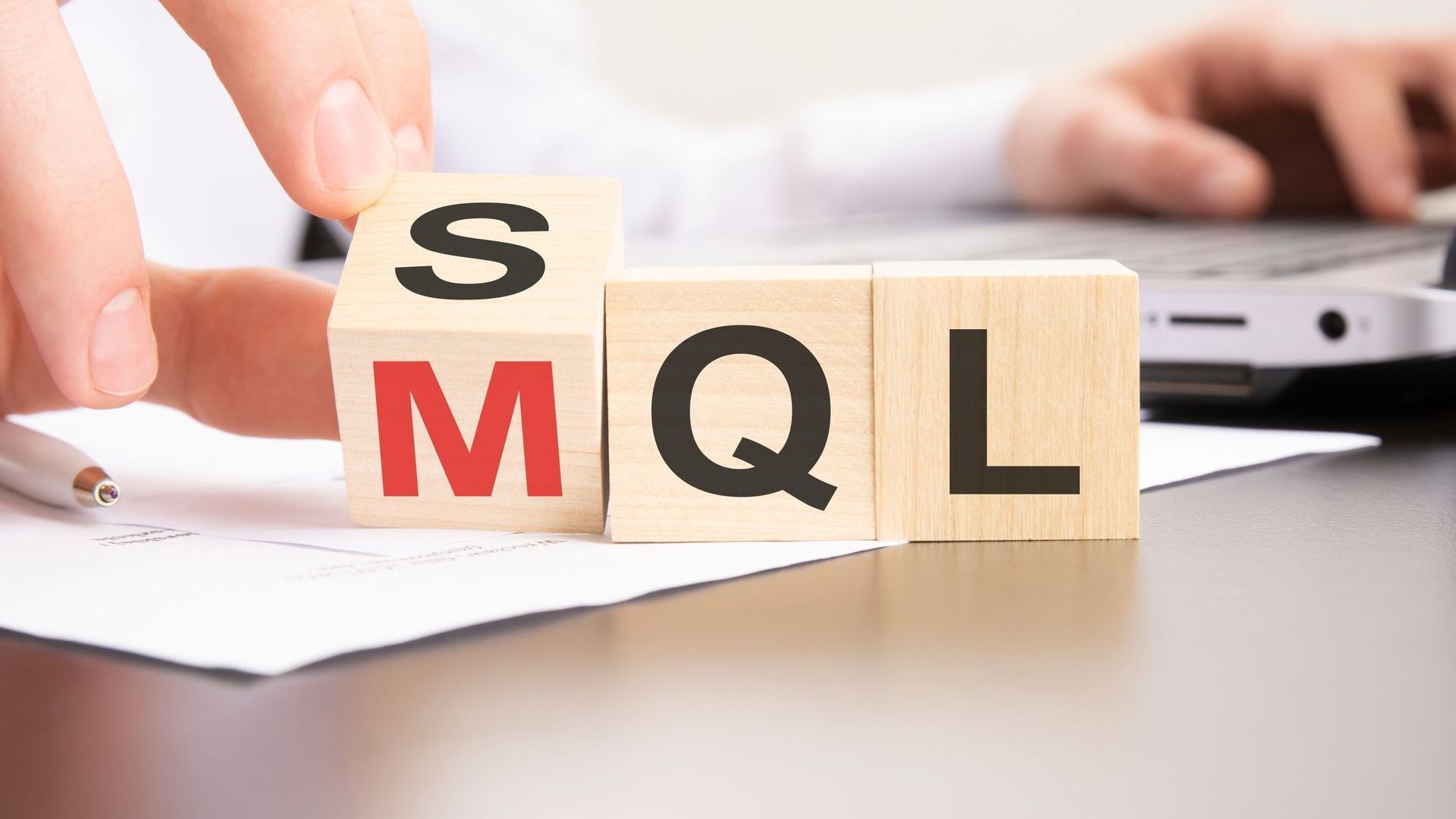 Wooden blocks spelling SQL on a desk with a hand arranging them near a laptop