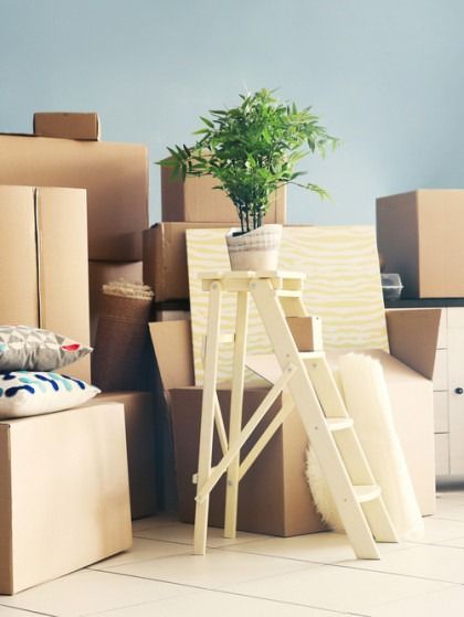 Cardboard boxes stacked for moving, small plant on a step stool, pillows, and rolled up padding.
