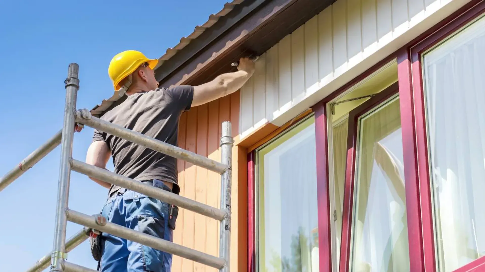 A person in blue coveralls and a cap uses a roller to paint a gray wall with peach-colored paint near a red stepladder.
