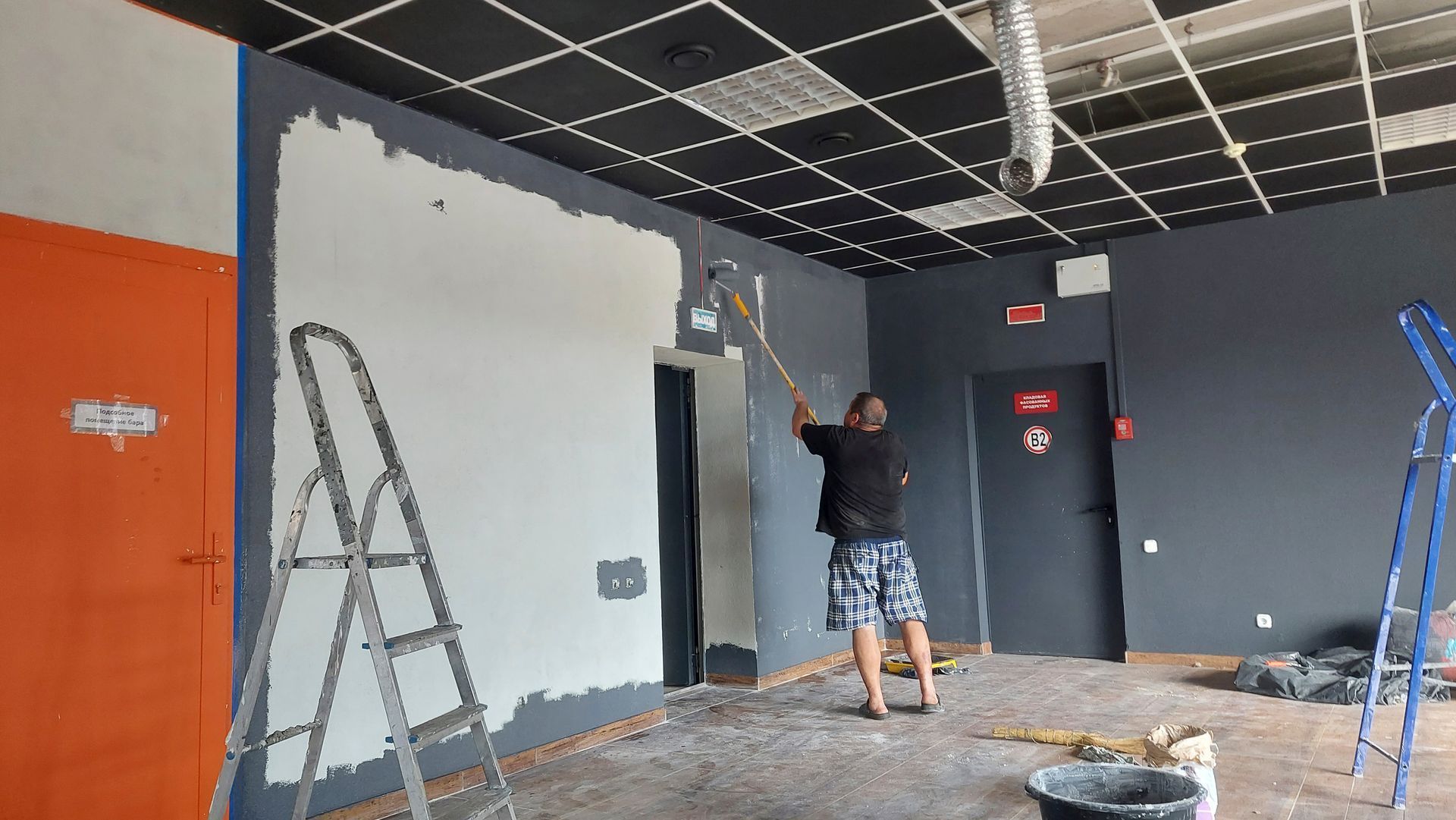 A person in shorts uses a long-handled tool to apply plaster to a dark gray wall in an unfinished room.