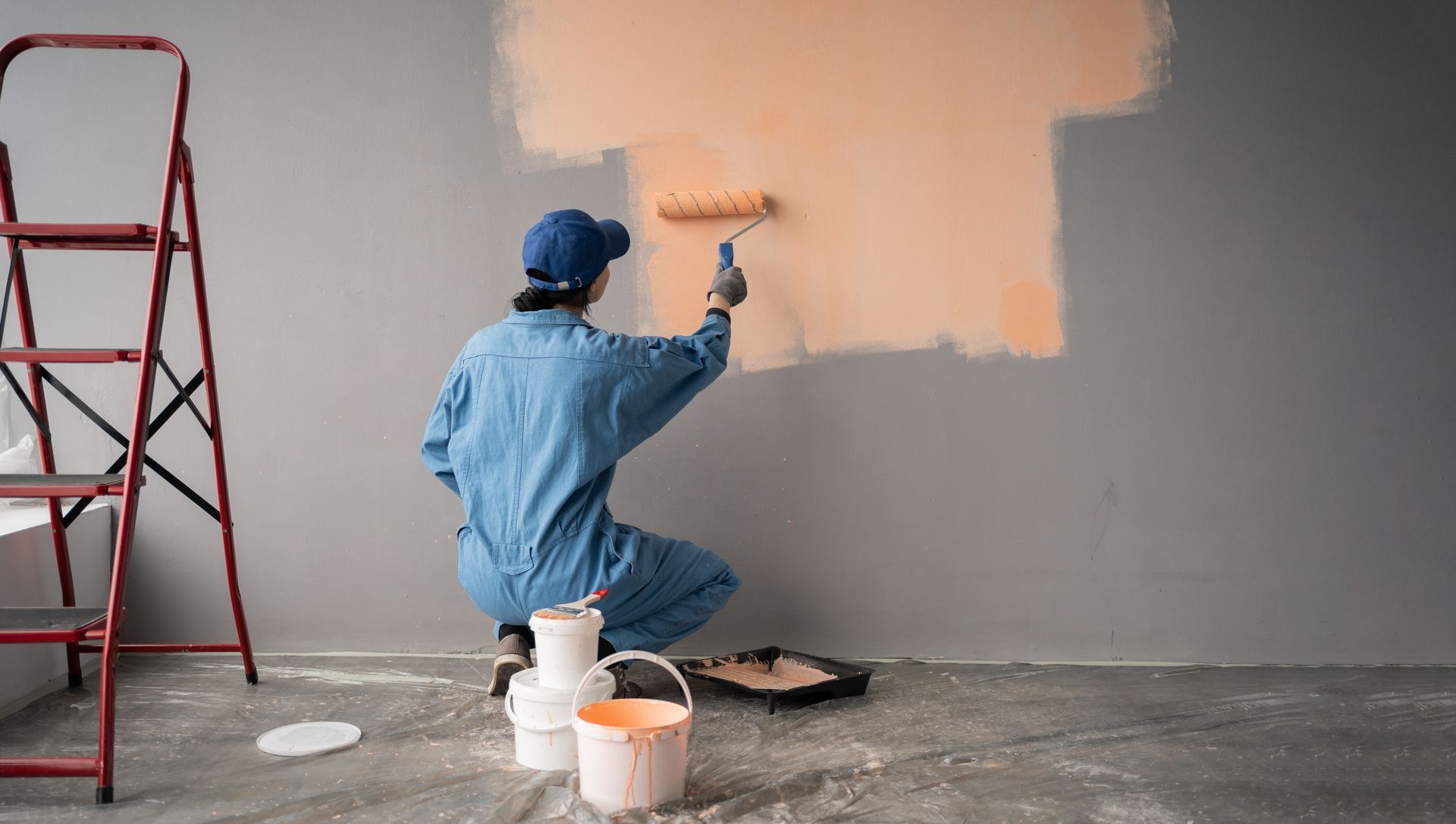 A worker in a green shirt and red overalls paints a white building wall while standing on scaffolding.