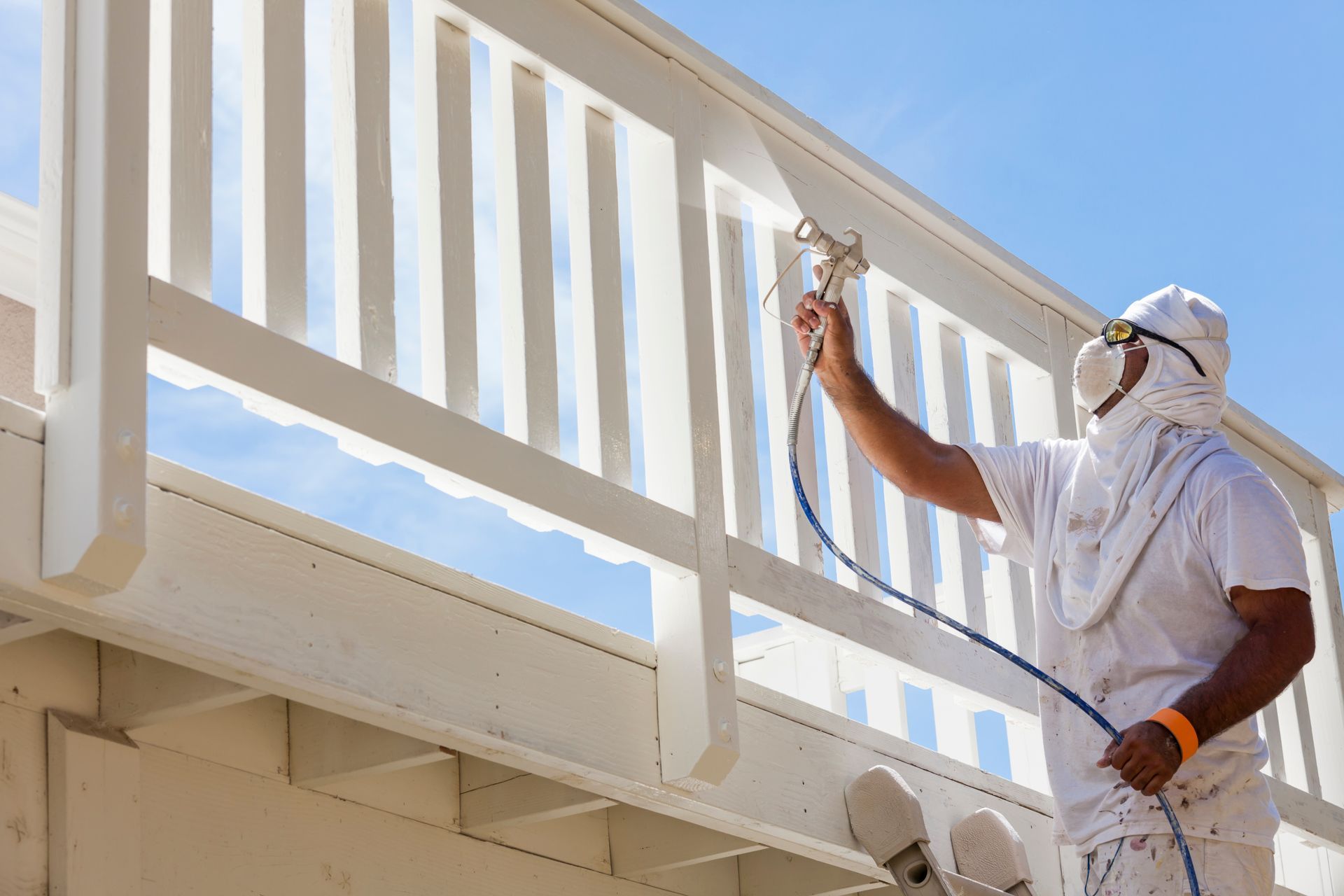 A painter in protective white gear and a respirator uses a spray gun to apply white paint to a wooden deck railing.