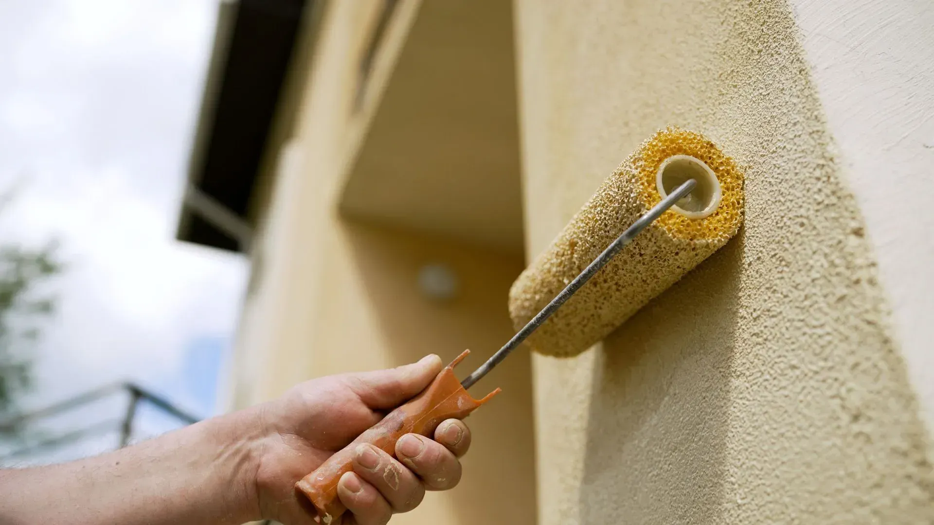 A person uses a yellow textured paint roller to apply light-colored paint to a rough exterior wall.